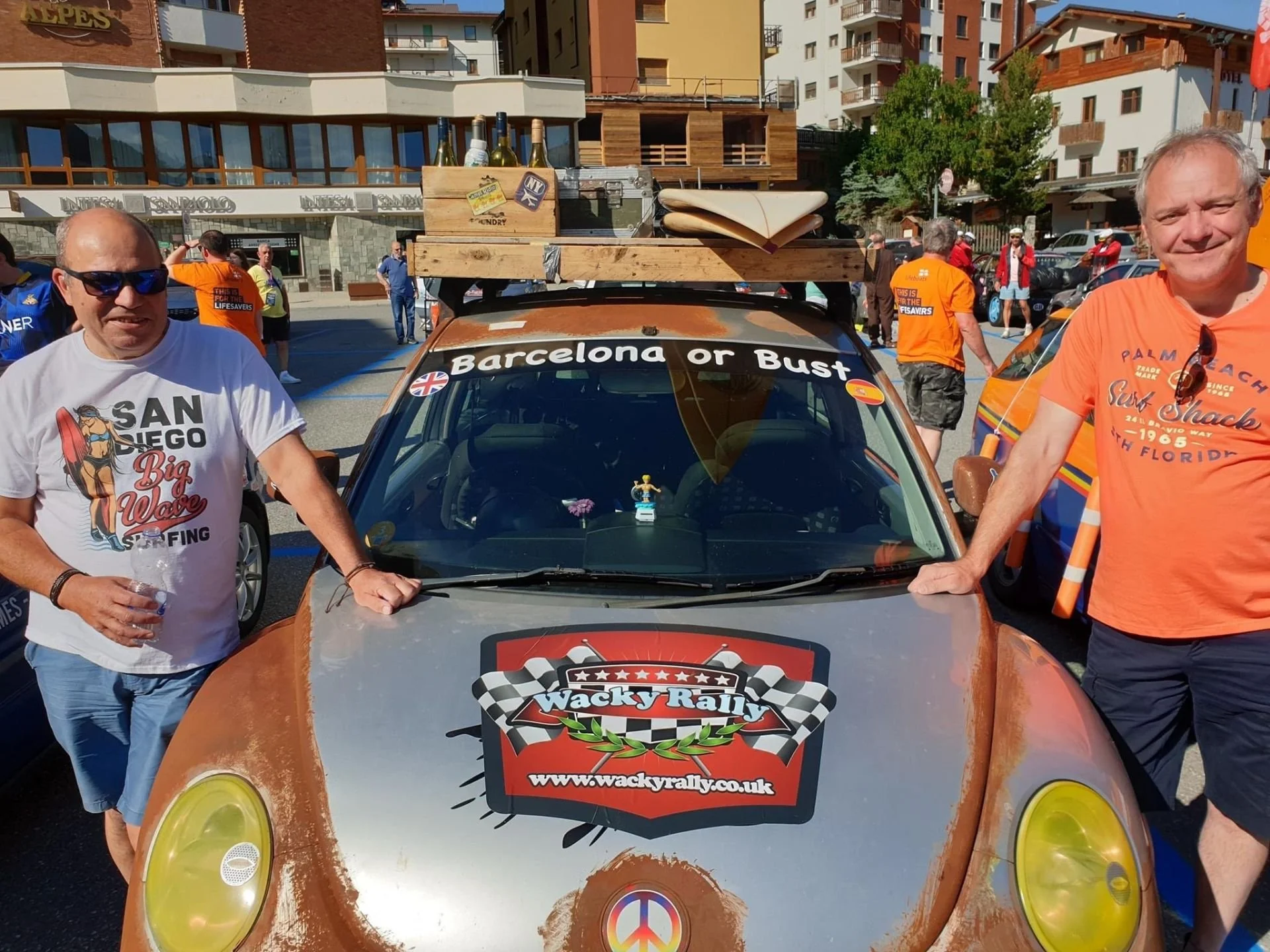 Two men standing beside a rally car in a parking lot during a rally event, with additional cars and people in the background. The car has rally stickers and a roof rack with bottles and supplies.