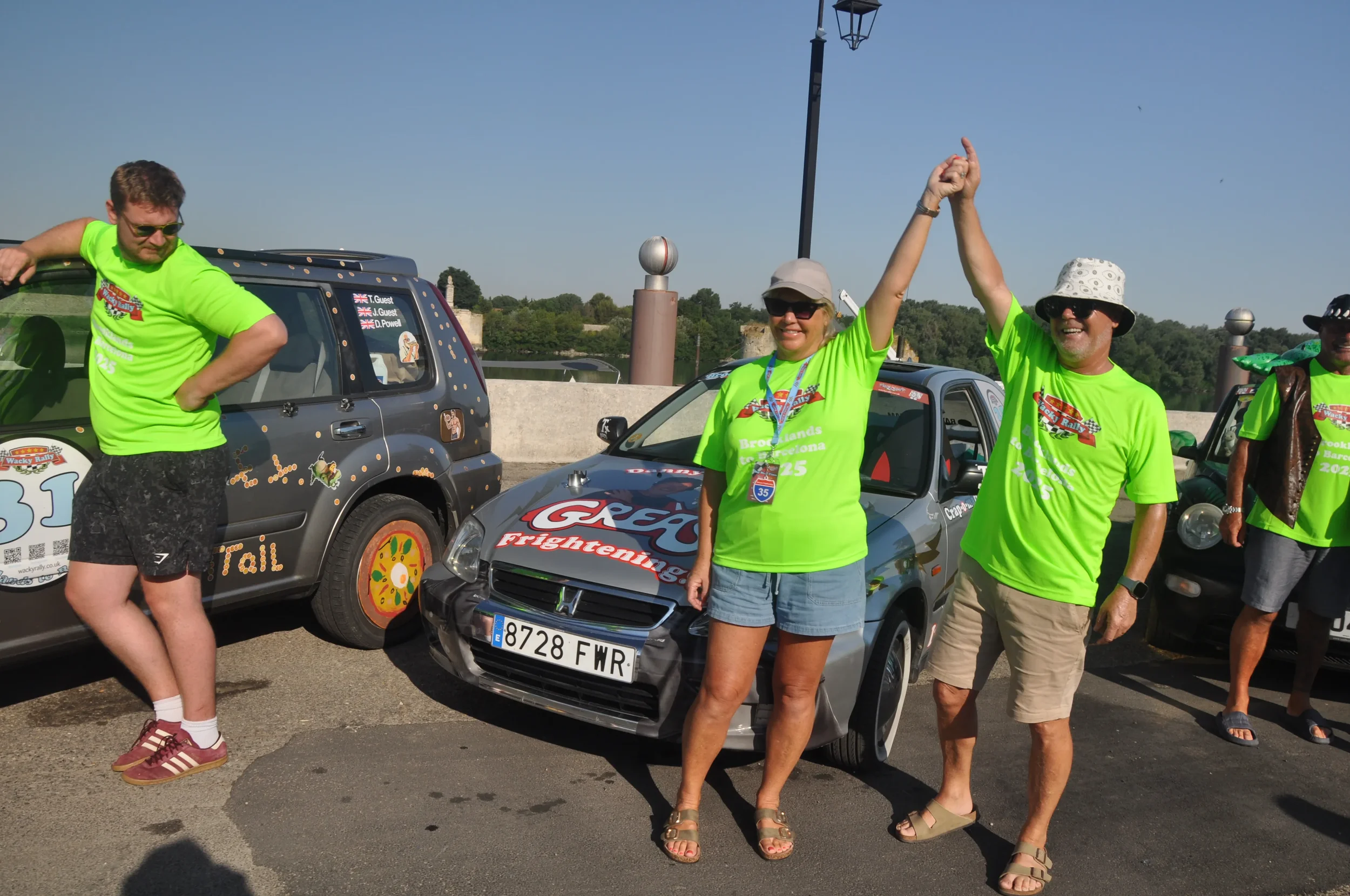 Group of people celebrating with cars in the background on a sunny day at a rally event. Two individuals in neon green shirts are holding hands in the center, smiling, one wearing a hat and sunglasses, the other wearing a cap and sunglasses. There ar