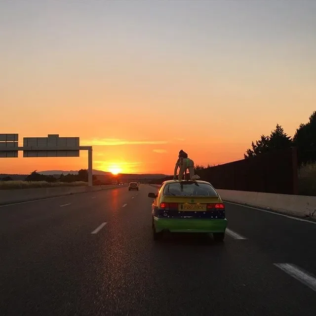 Person sitting on top of a yellow and green car on a highway during a sunset.
