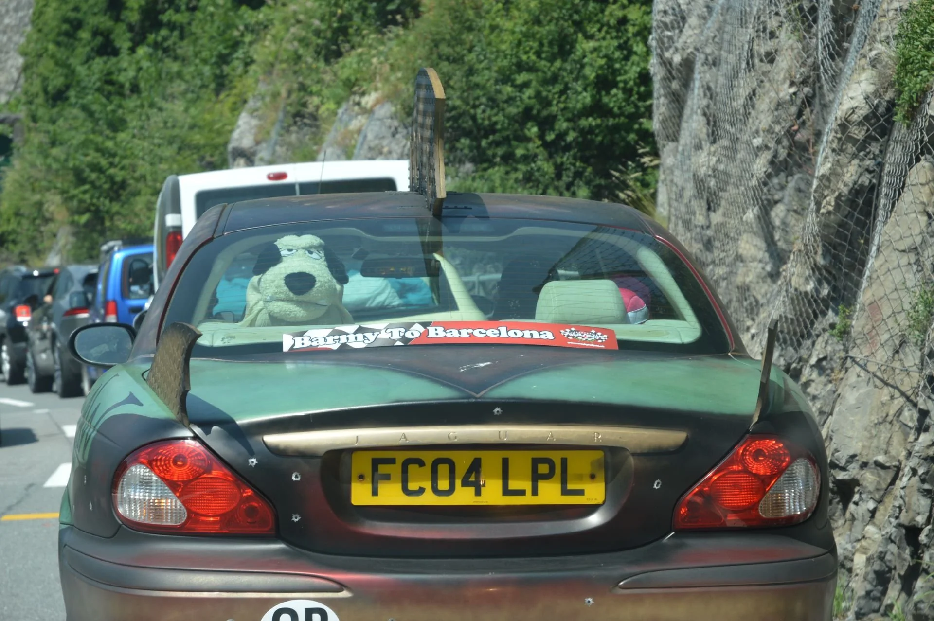 A line of cars on a mountain road with a Jaguar convertible in front. The convertible has a stuffed dog toy in the back window and a bumper sticker that reads 'Barny To Barcelona.'