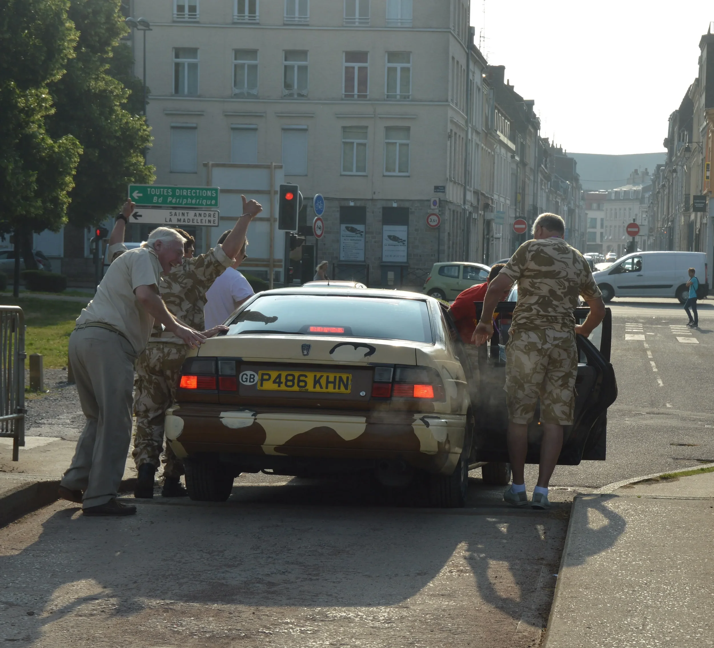 A group of people, some in camouflage clothing, gathered around a beige car with camouflage paint on a city street, appearing to be working on or inspecting the vehicle.