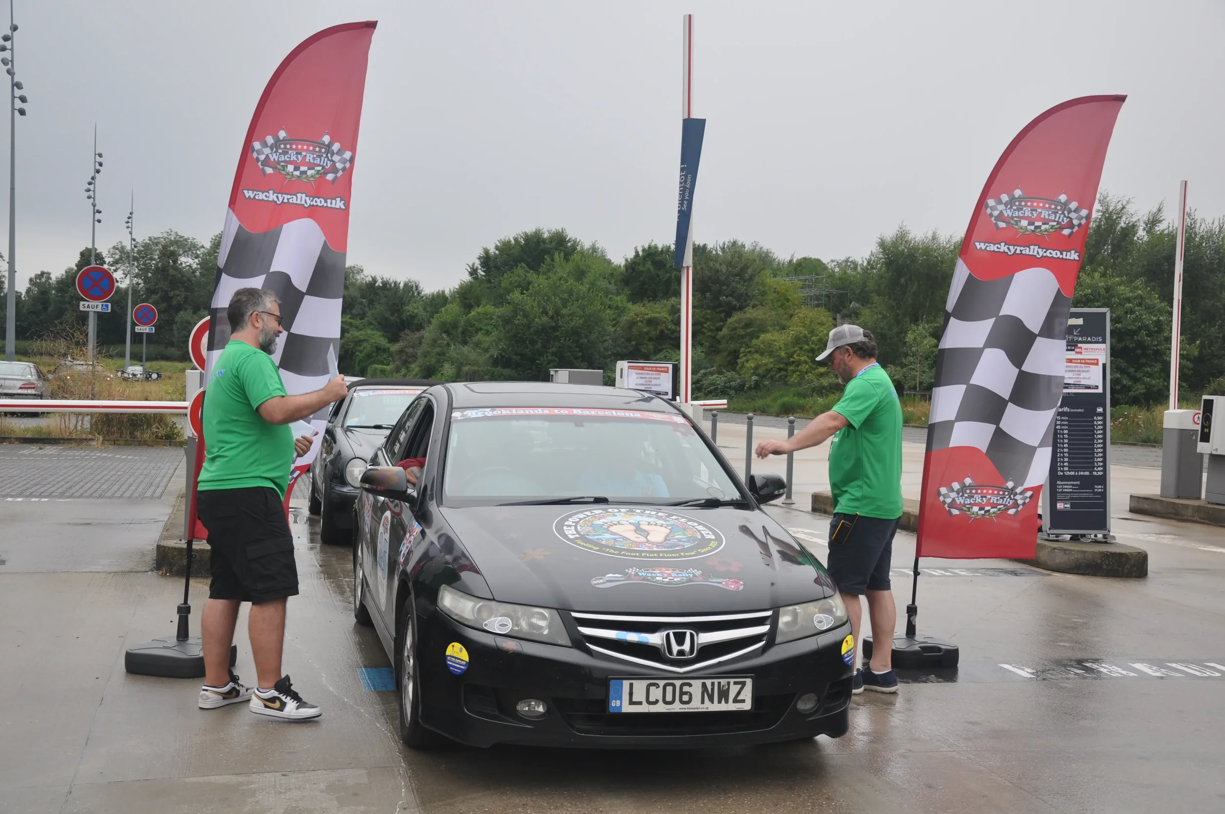 Two men in green shirts standing next to a black Honda car at a rally event, with flags and banners that read "Wacky Rally" in the background.
