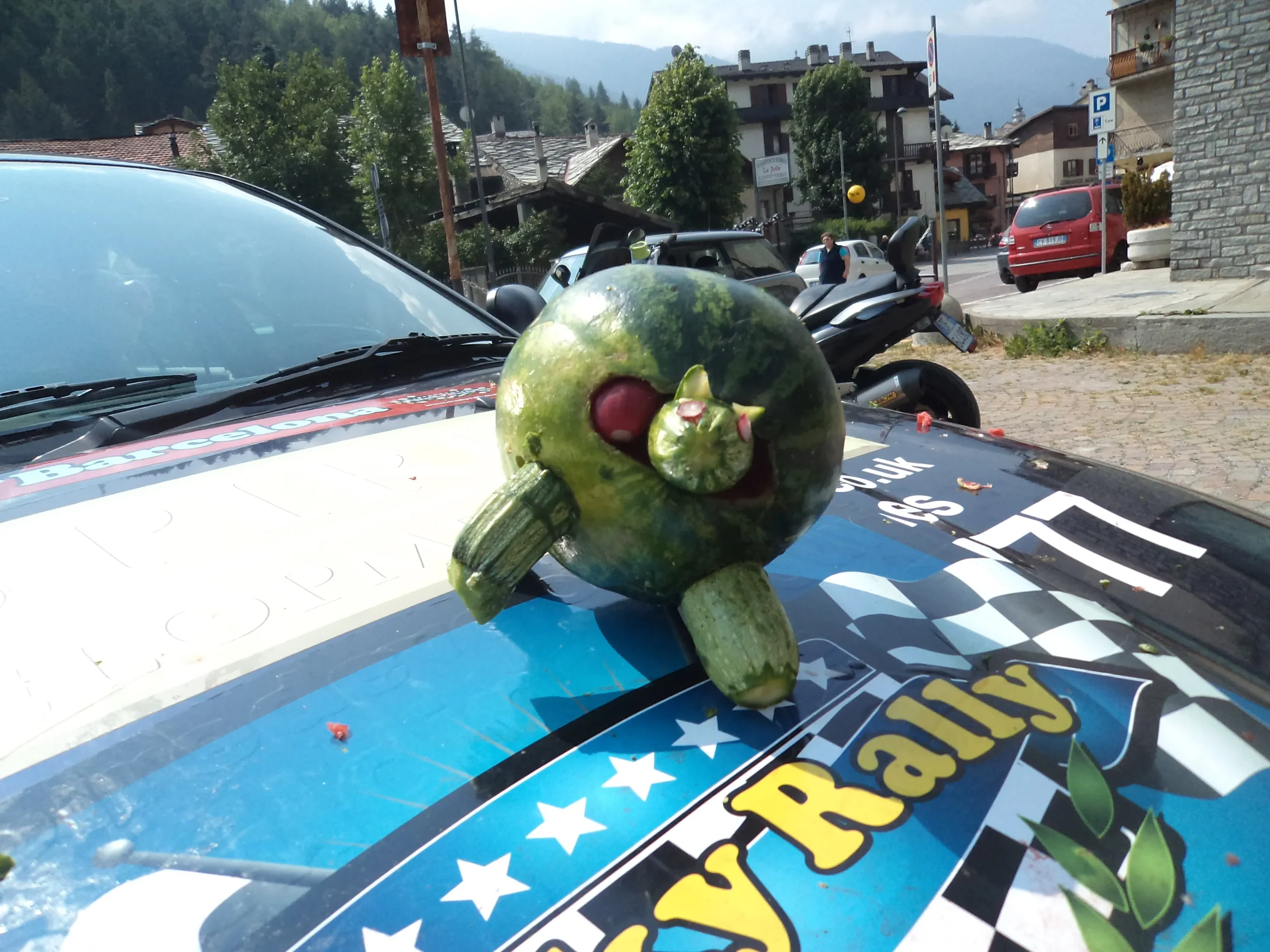 A watermelon decorated to look like a cartoon face, with a small watermelon used as a nose and a carved opening for a mouth, placed on top of a police car with a sticker that says 'Rally' and a checkered pattern, in an outdoor town setting with build