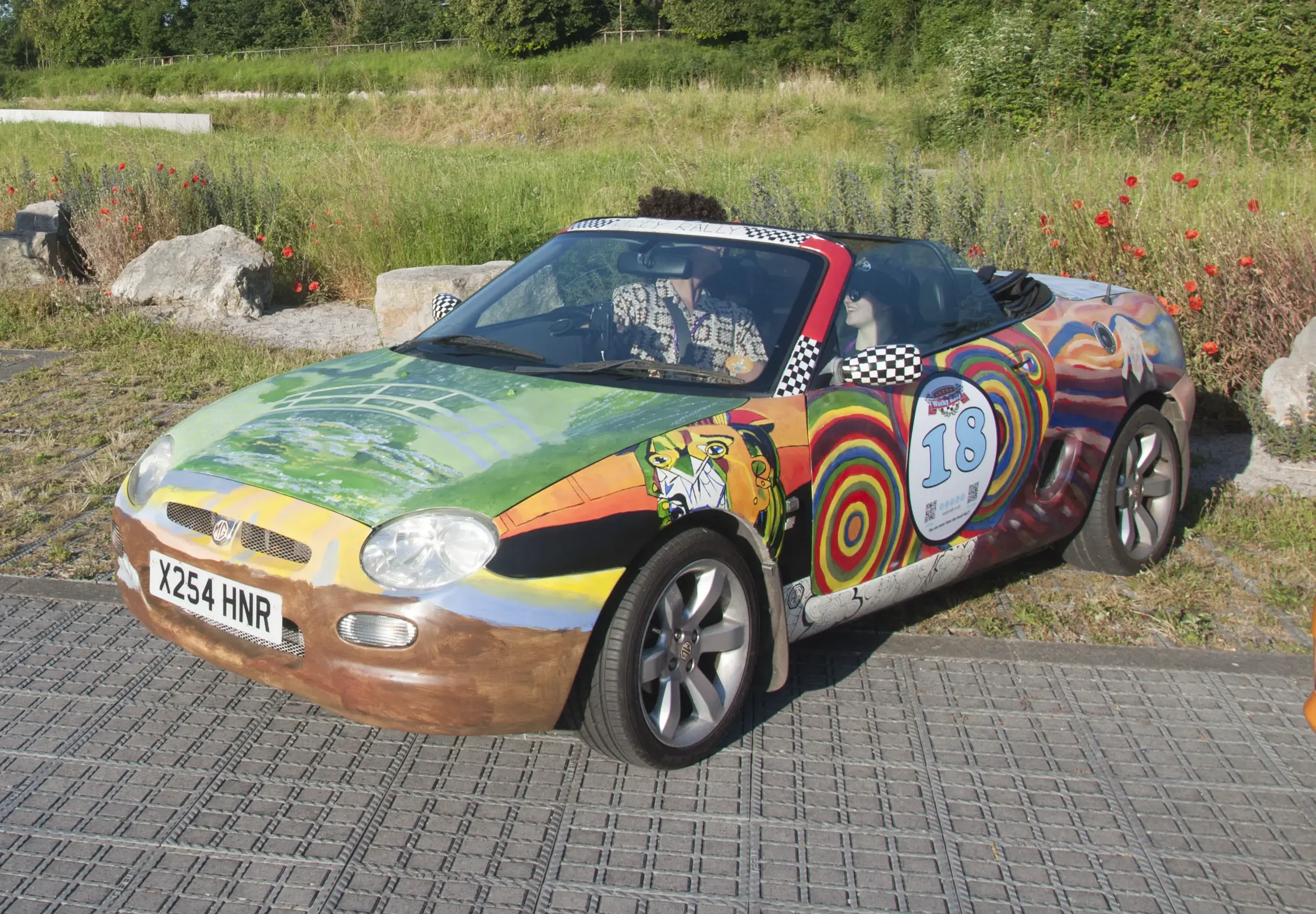 A colorful, artistic sports car with a diverse, vibrant mural design parked on a paved area near a grassy field with rocks and flowers.