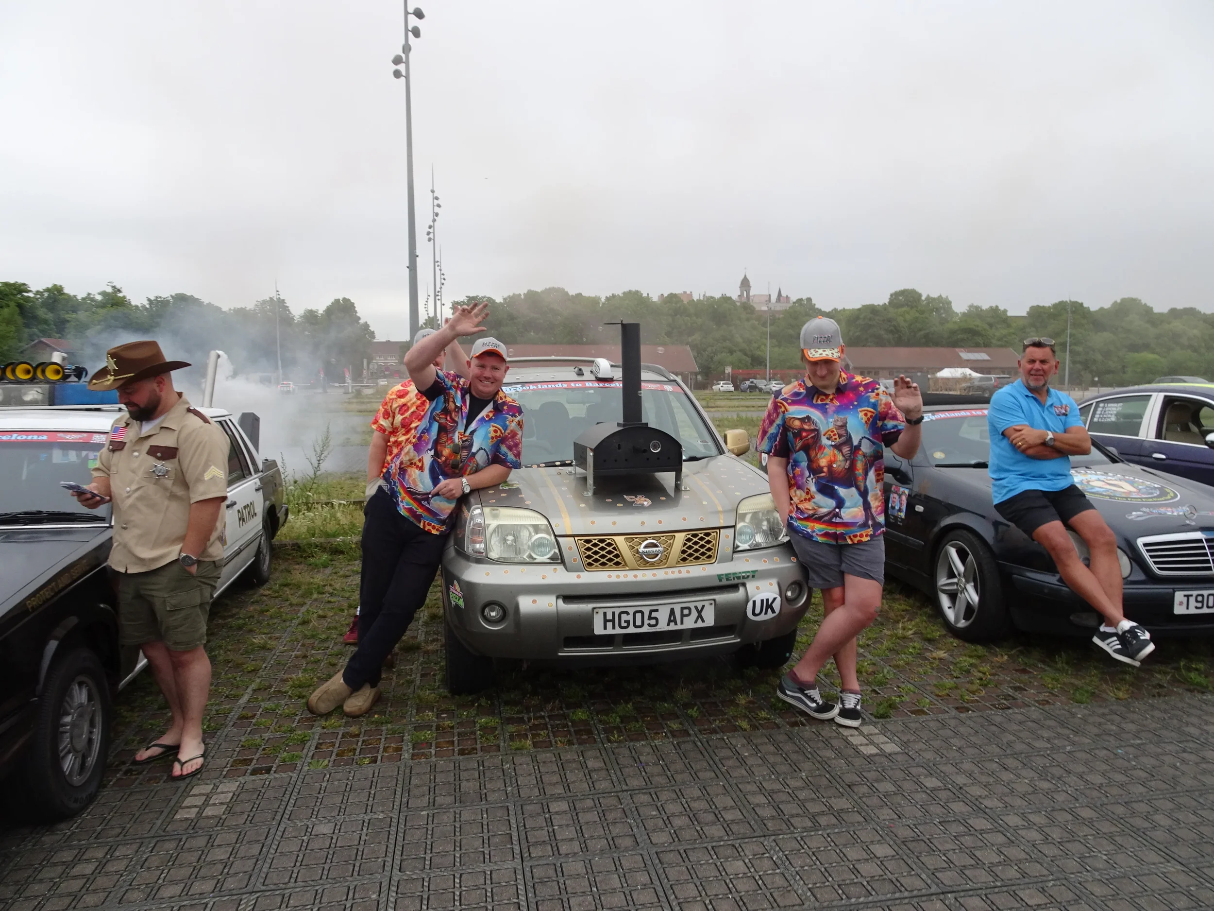 Four men standing in front of cars at an outdoor event: one in a uniform and three in colorful shirts, with two of them waving.