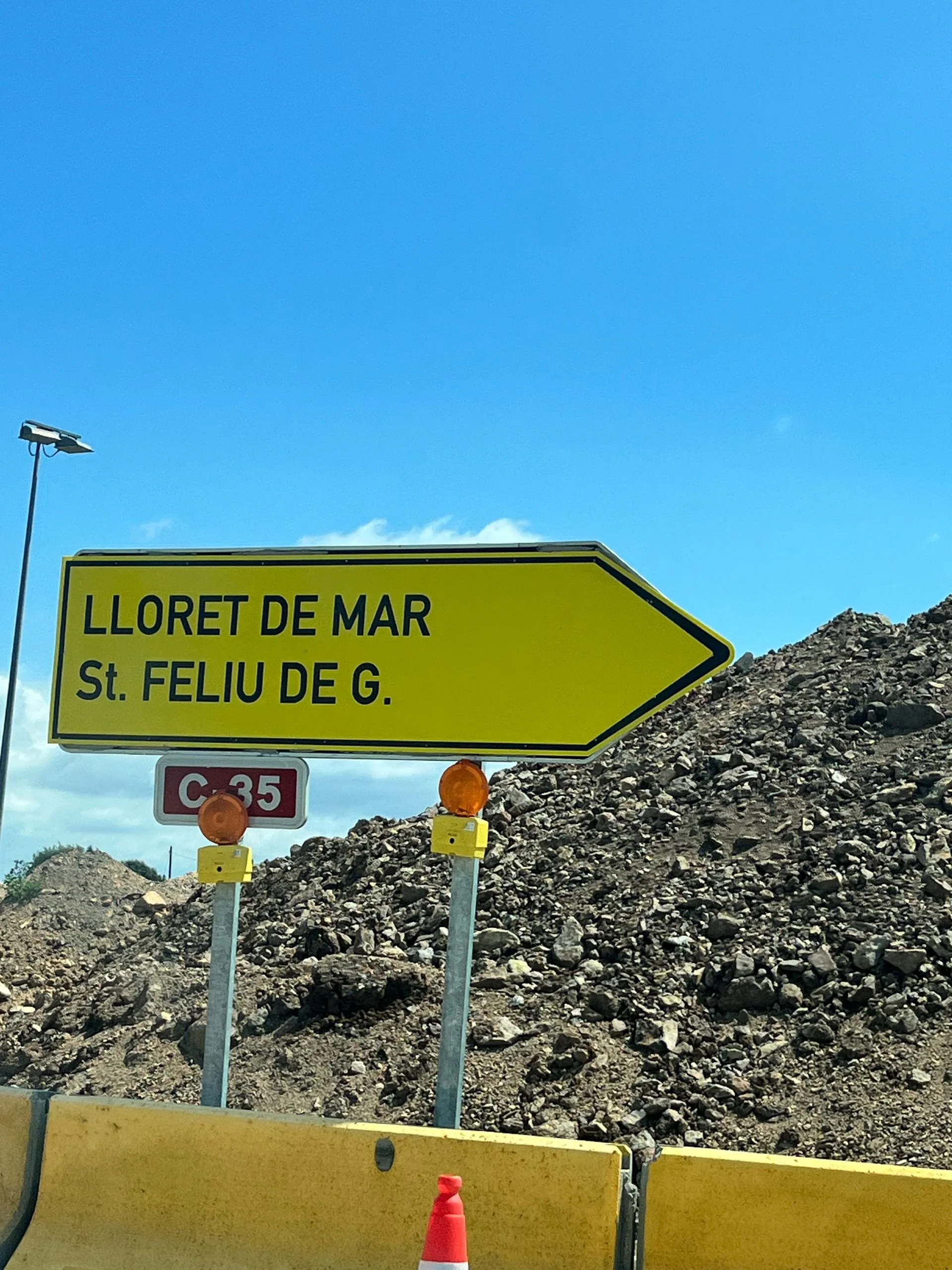Yellow road sign indicating directions to Lloret de Mar and St. Feliu de G. under a blue sky, with pile of rocks and construction barriers and traffic cones in the foreground.
