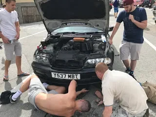 Three men working under the open hood of a black car parked in an outdoor lot, with one man lying on the ground and two others kneeling nearby.