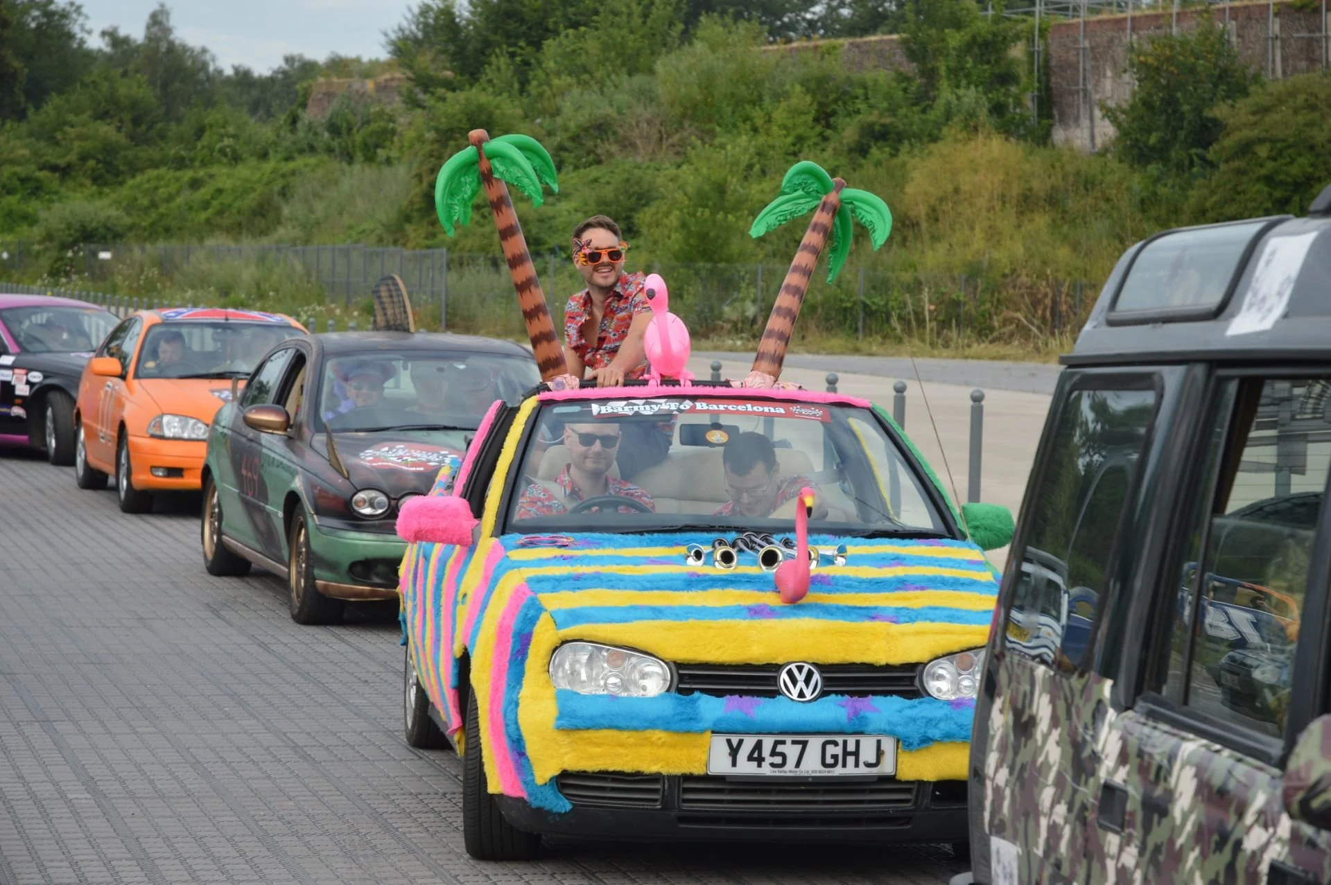 Colorful decorated car with inflatable palm trees, pink flamingo, and googly eyes on the front, driving on a street with other cars; people inside wearing colorful shirts, one standing through the sunroof wearing sunglasses.