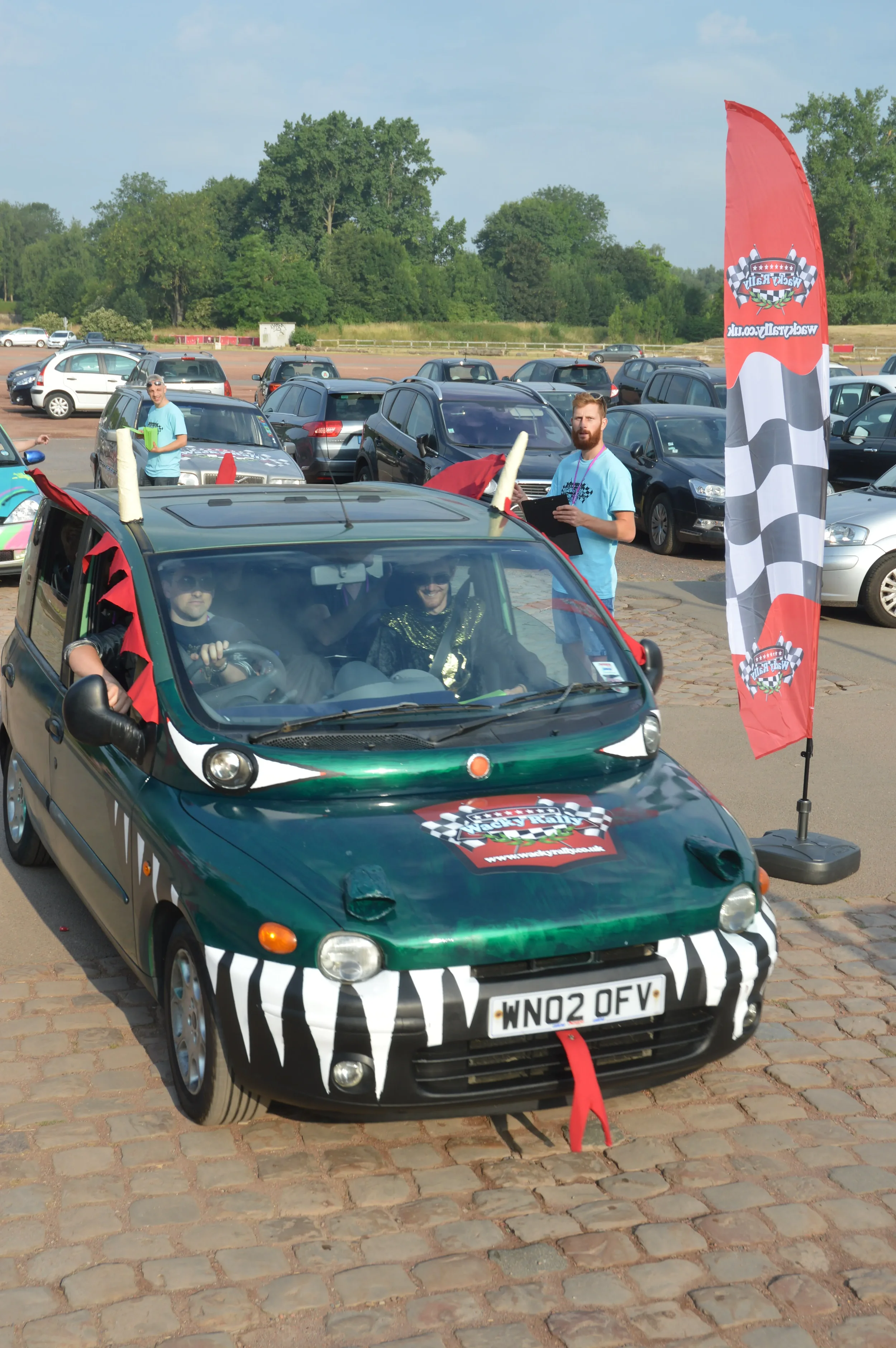 A small car decorated with zebra stripe patterns and a racing logo, with two people dressed in zebra-like costumes, participating in a fun event or parade at a parking lot, with a red flag and people standing around.