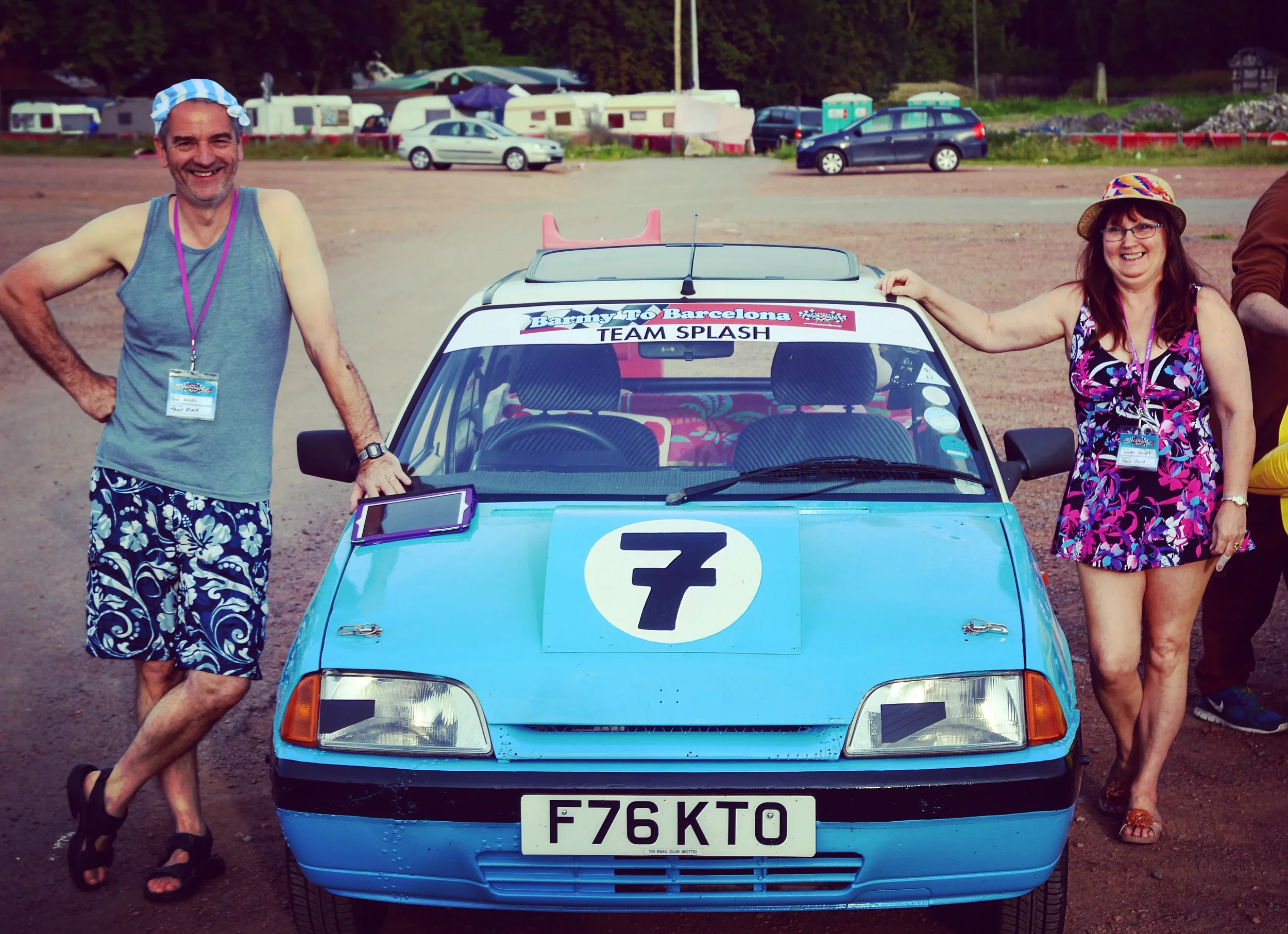 Two smiling people, a man and a woman, stand beside a small blue rally car with the number 7 on the hood, in a parking lot with caravans in the background. The man wears a blue cap, gray tank top, and floral shorts, while the woman wears a floral dre