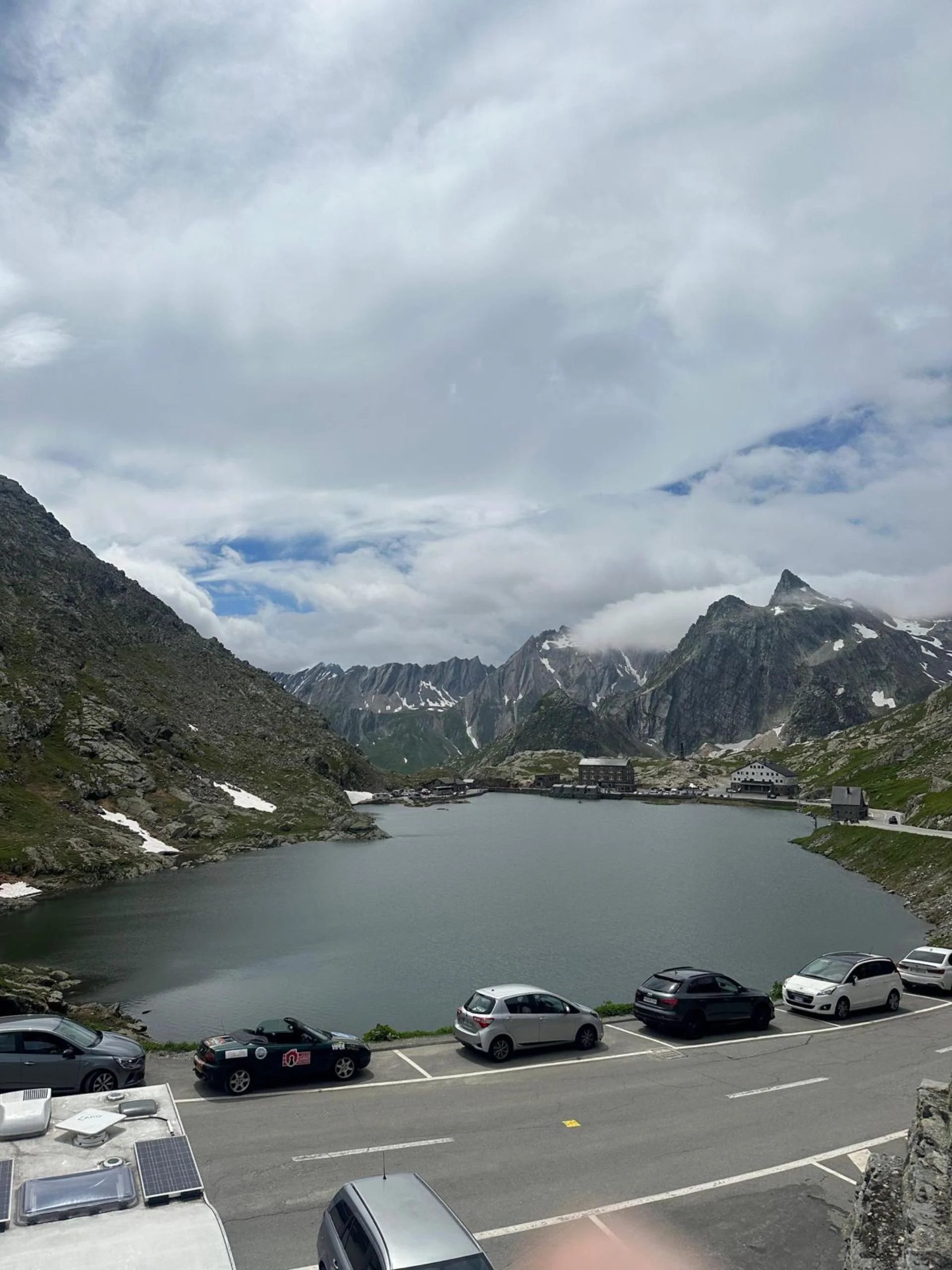 Mountain lake with surrounding mountains, parked cars in foreground, cloudy sky overhead.