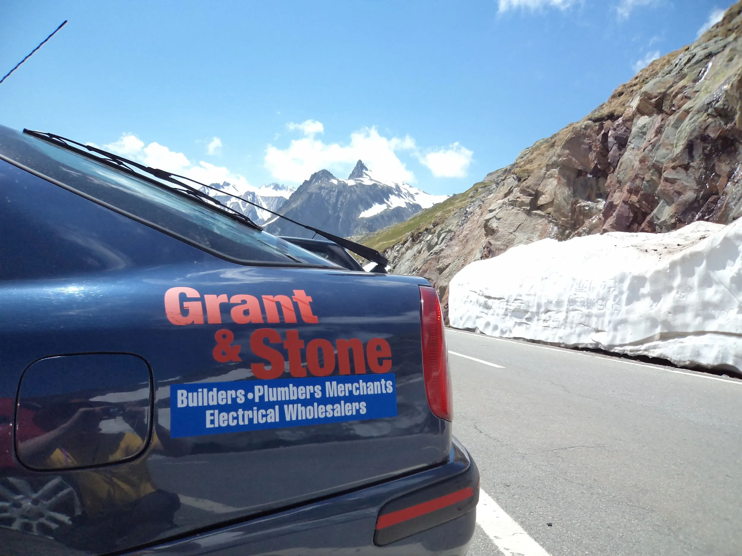Close-up of a vehicle with a sign reading 'Grant & Stone' and a list of services, parked on a mountain road with snow and rocky terrain, snow-capped mountains in the background, and a blue sky with clouds.