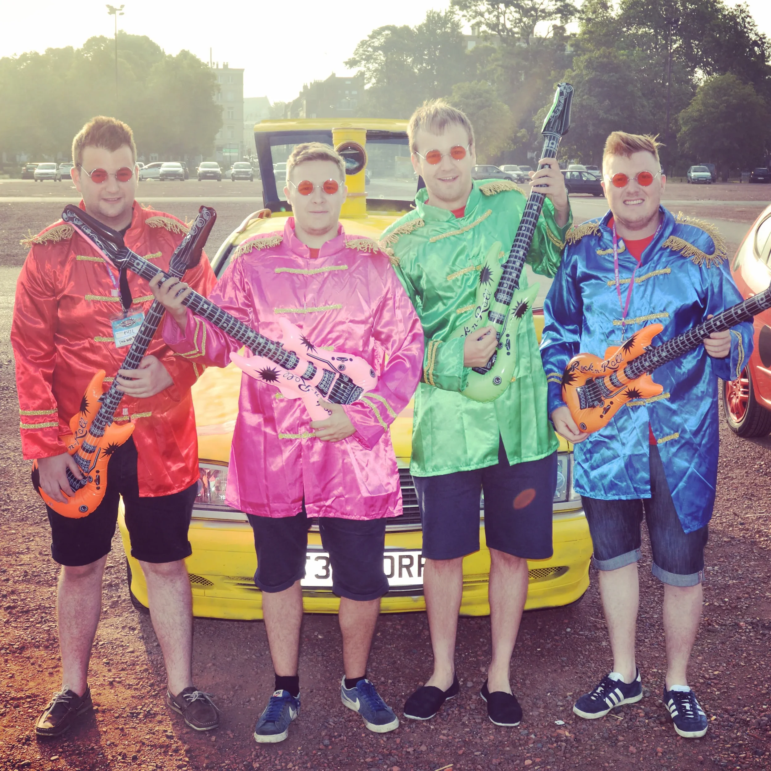 Four men dressed in colorful, shiny rock 'n' roll costumes holding inflatable guitars in front of a yellow car, with a park and city buildings in the background.