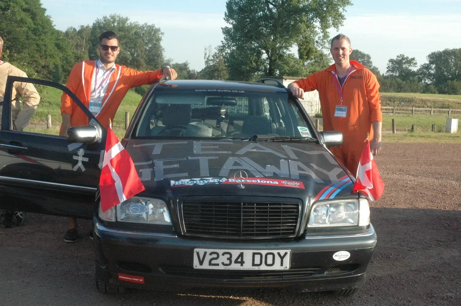 Two smiling men in orange coveralls standing beside a black rally car with Danish flags, outdoors with trees and a field in the background.