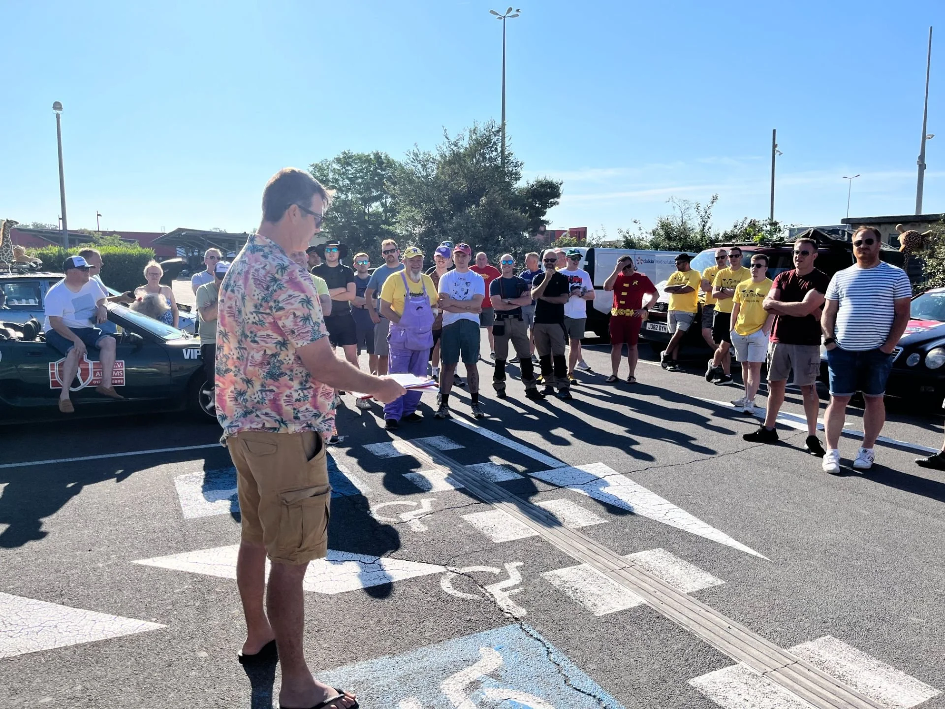 A man in a floral shirt and beige shorts is reading or giving a speech to a crowd gathered in a parking lot. The crowd includes people dressed casually, some wearing yellow jerseys or t-shirts, and one person in a purple outfit. Some cars are parked 