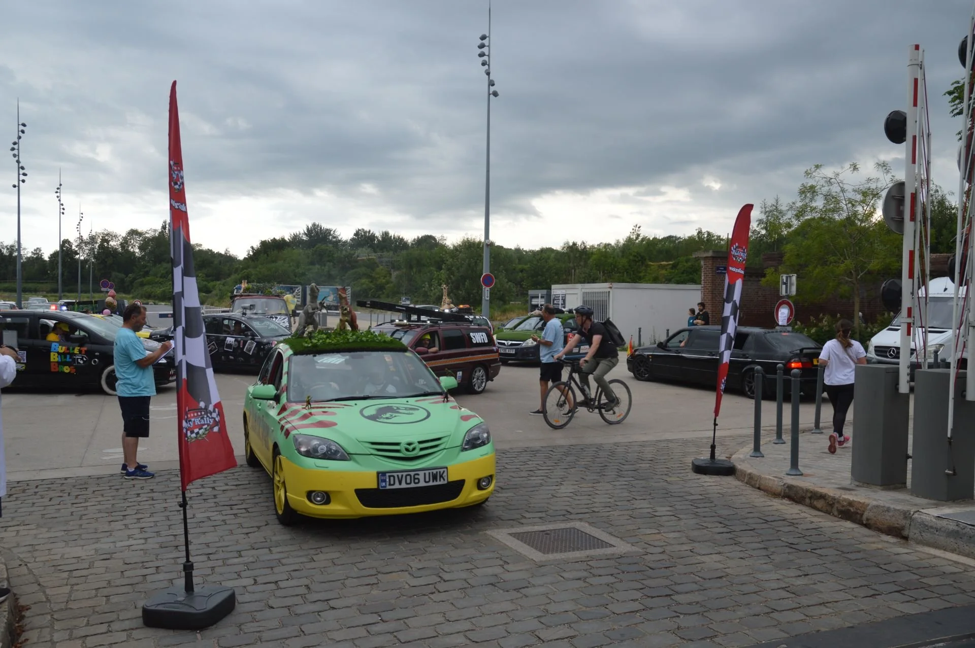 A green and yellow Mazda car with car decorations on the roof, parked among other cars, with flags and people around, including a person riding a bicycle and another checking their phone. The scene is outdoors on a cloudy day.