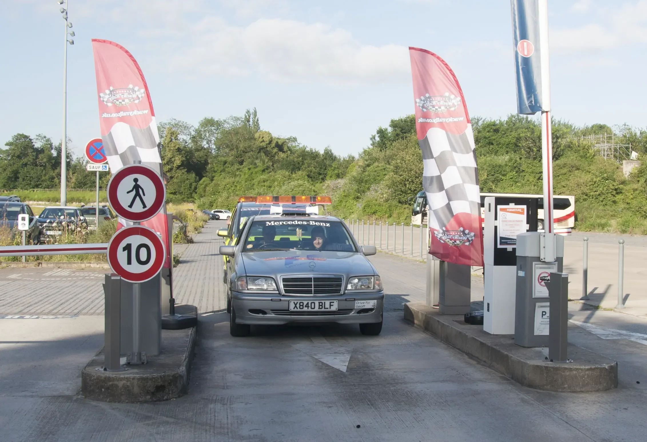 A Mercedes-Benz car passing through a parking gate with red banners on either side featuring checkered flags, a speed limit sign of 10, and a walking person sign, in a parking lot with trees and other vehicles in the background.