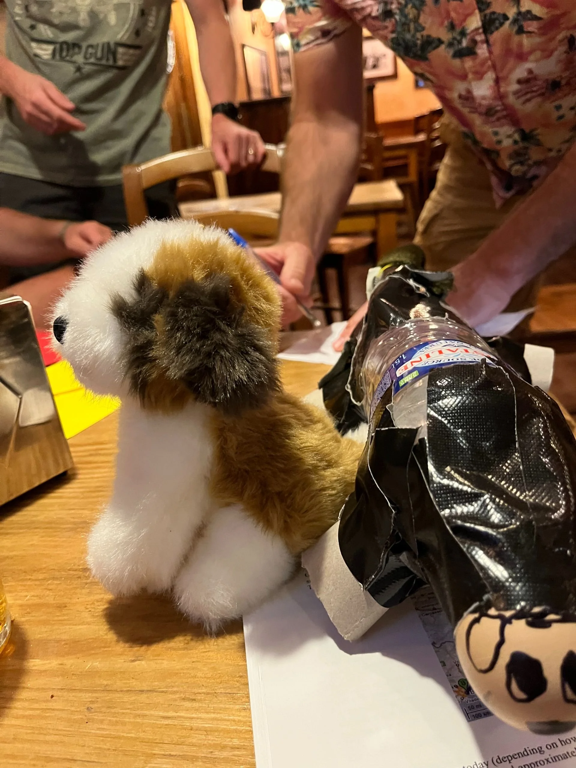 A stuffed animal resembling a rabbit with brown, black, and white fur sitting on a table, with people around, in a casual indoor setting.