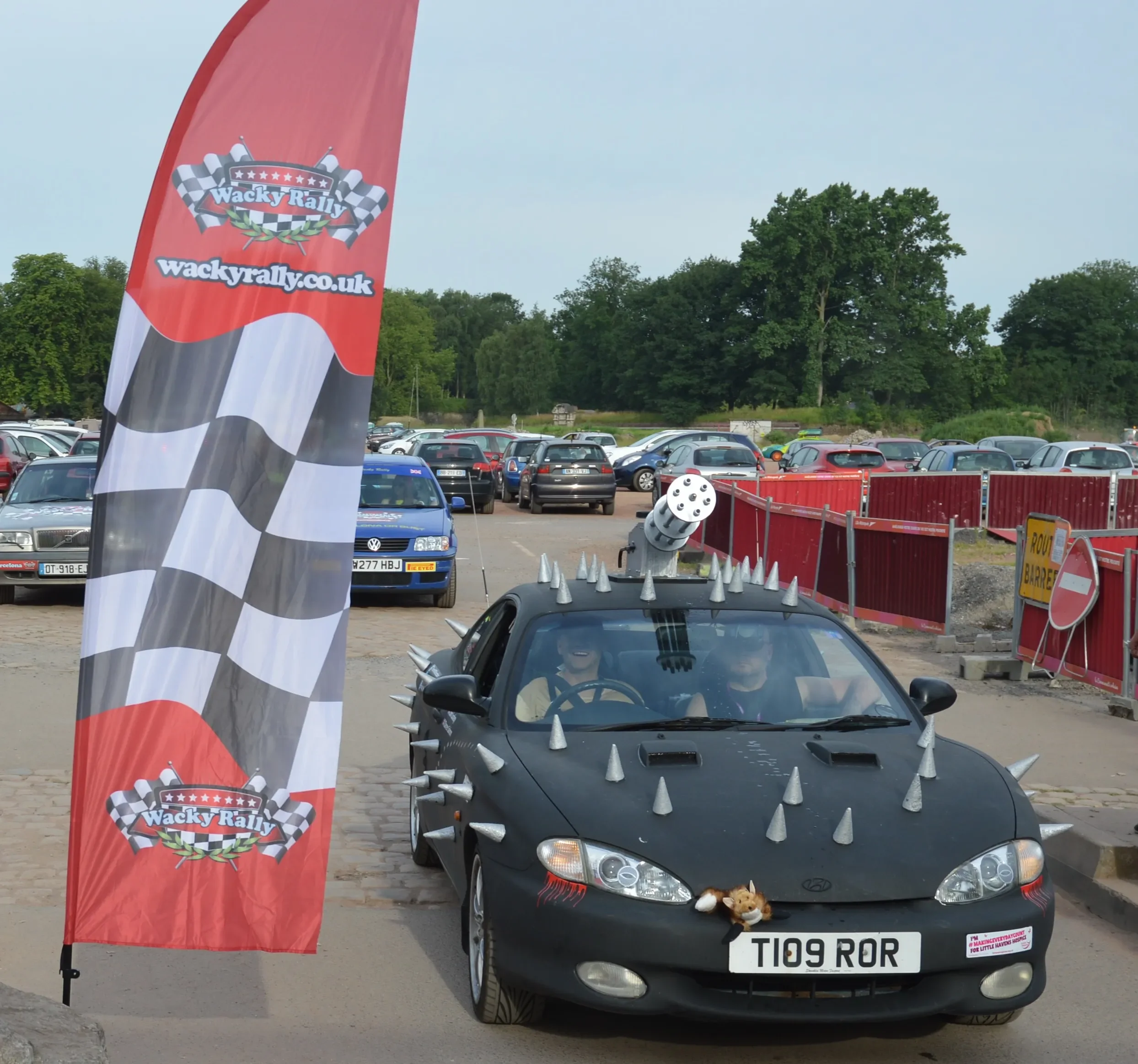 A black sports car decorated with silver spikes, with a toy lion hanging from the front grille, parked near a red flag with checkered flags andquot;Wacky Rallyandquot; and a website URL. The car has a mounted mock missile on the roof, and two people 