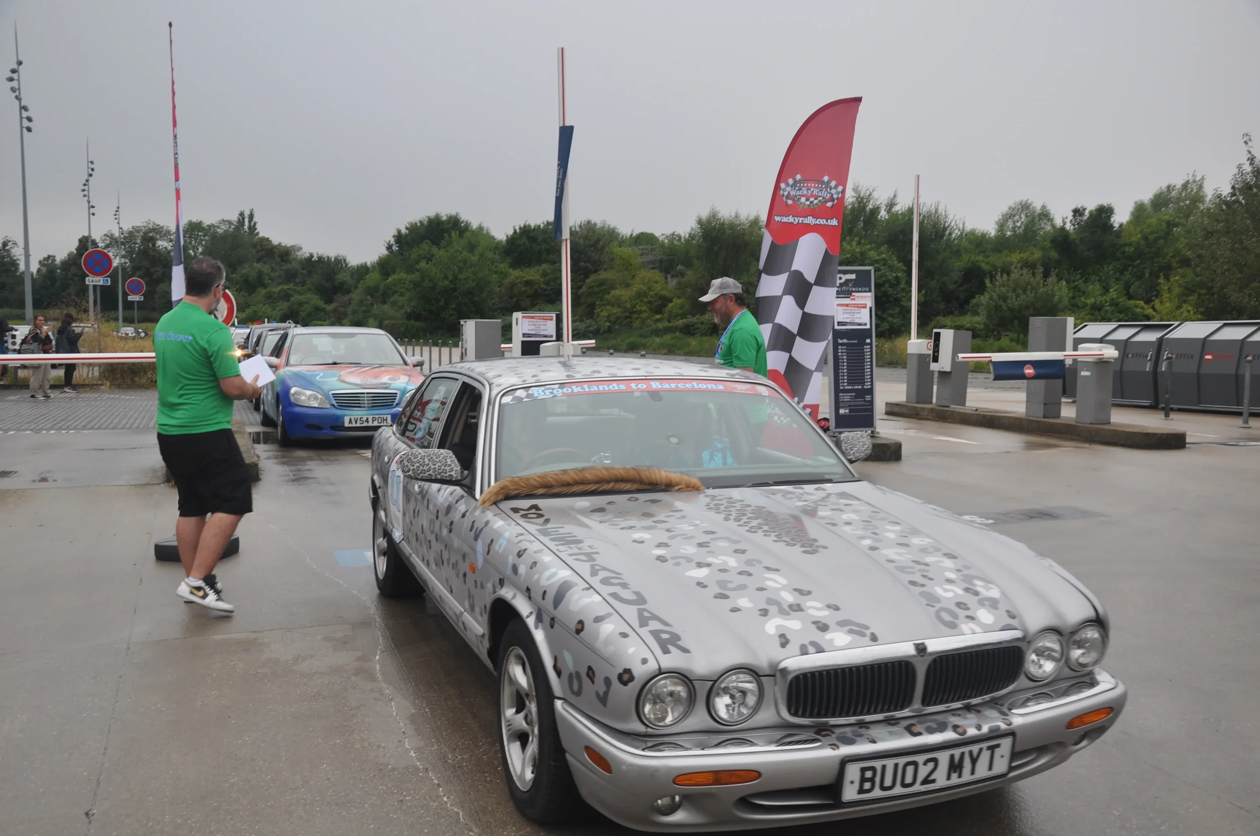 A vintage silver Jaguar XJ-S with leopard print details parked at a race event with people in green shirts, a checkered flag banner, and other race cars in the background.