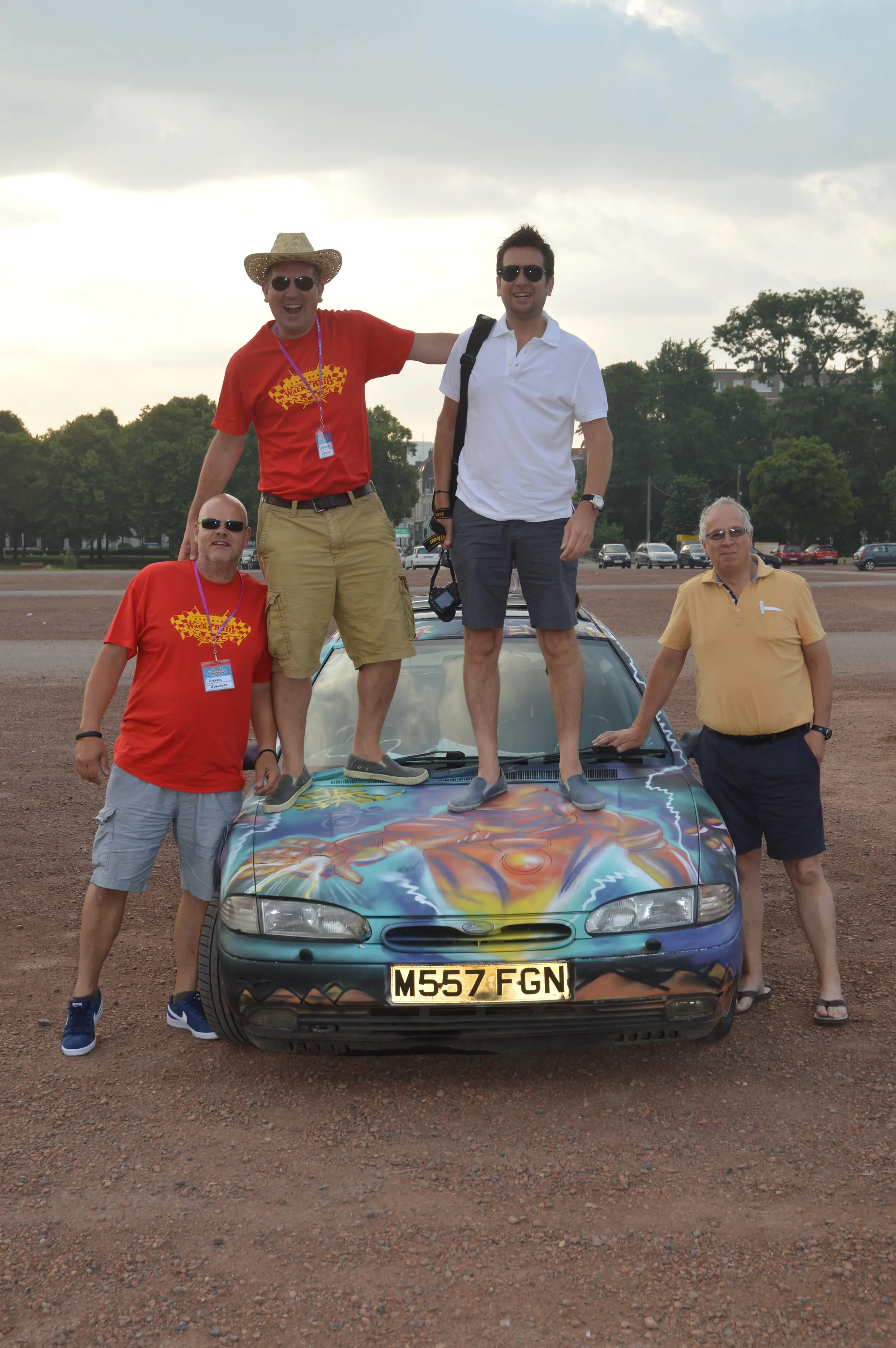 Four men posing on and around a colorful, custom painted car in an outdoor parking lot during the day. Two men are standing on the car's hood, one on the ground to the left and the other to the right. Two men are standing beside the car, one on the h