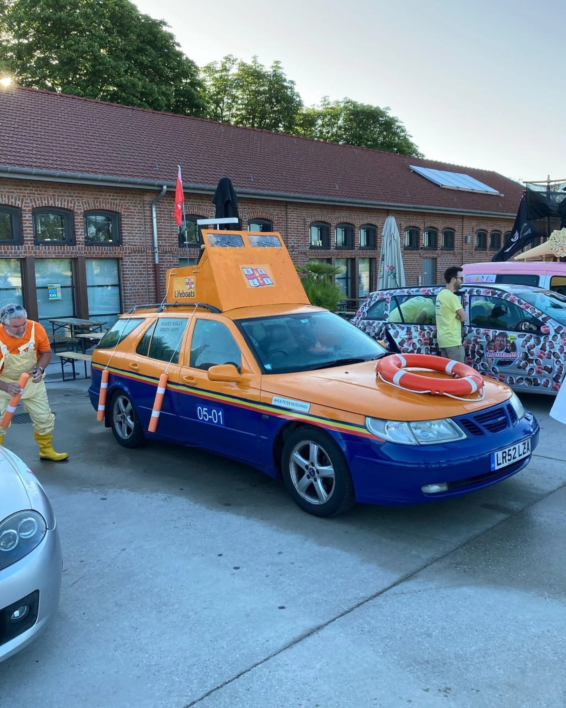 A car decorated to resemble a lifeboat, with orange and blue colors, a life ring on the hood, and a sign indicating 'Lifeboats.' It has a station on top with windows and a flag. The car is parked outdoors with other vehicles and people nearby.