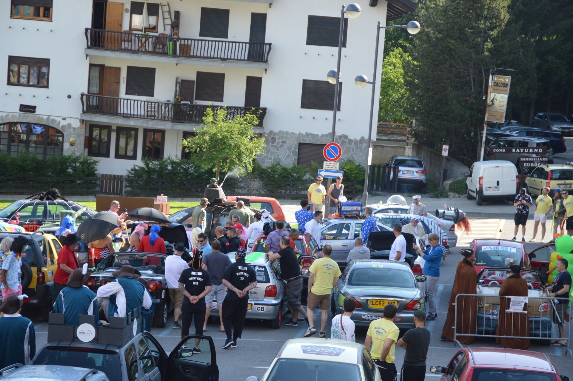 A lively outdoor festival with numerous costumed people, vintage cars, and spectators gathered in a street near a white building with balconies, trees, and parked vehicles.
