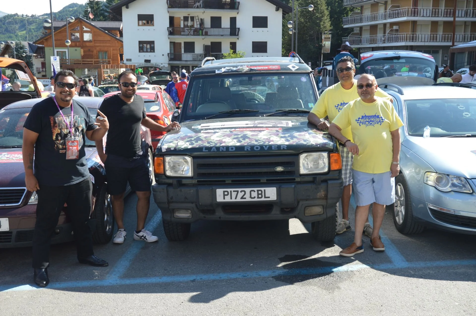 Group of five men standing in front of a black Land Rover, at a car gathering or event, with a background of buildings and parked cars.