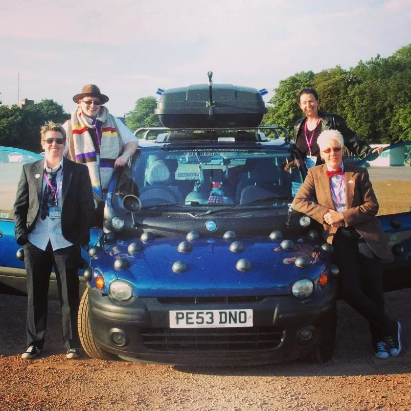 Four people standing around a blue car decorated with numerous spherical objects. The car has a roof cargo box, and the background shows outdoor scenery with trees.