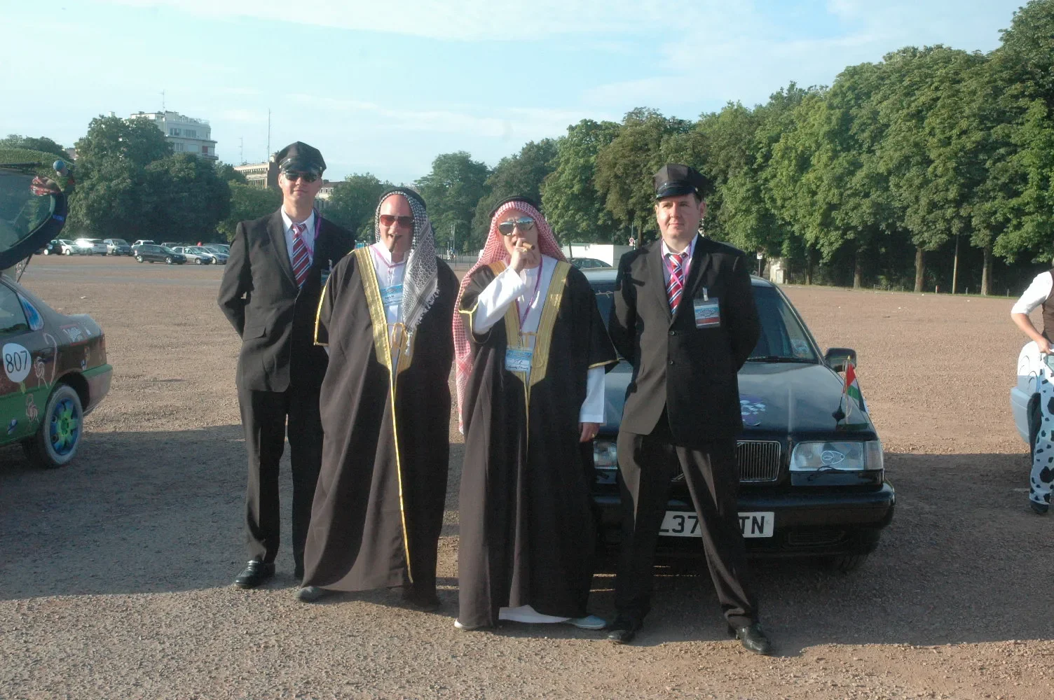 Four people dressed in costumes, with two wearing what appear to be Middle Eastern attire and the other two in black suits with ties, standing in front of a black car in an outdoor parking lot with trees in the background.