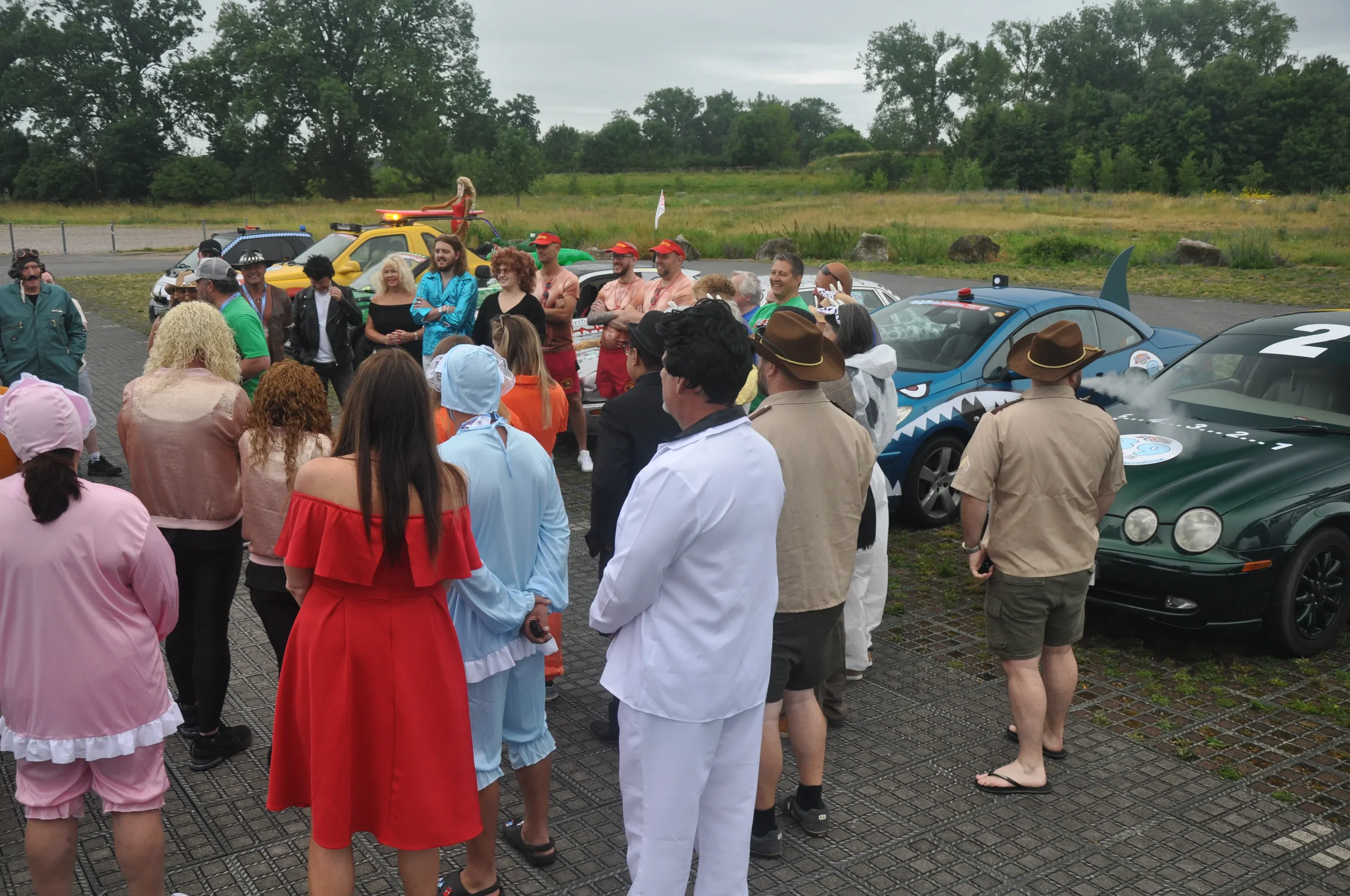 Group of people dressed in various costumes gathered outdoors near decorated cars in a parking lot during daytime.