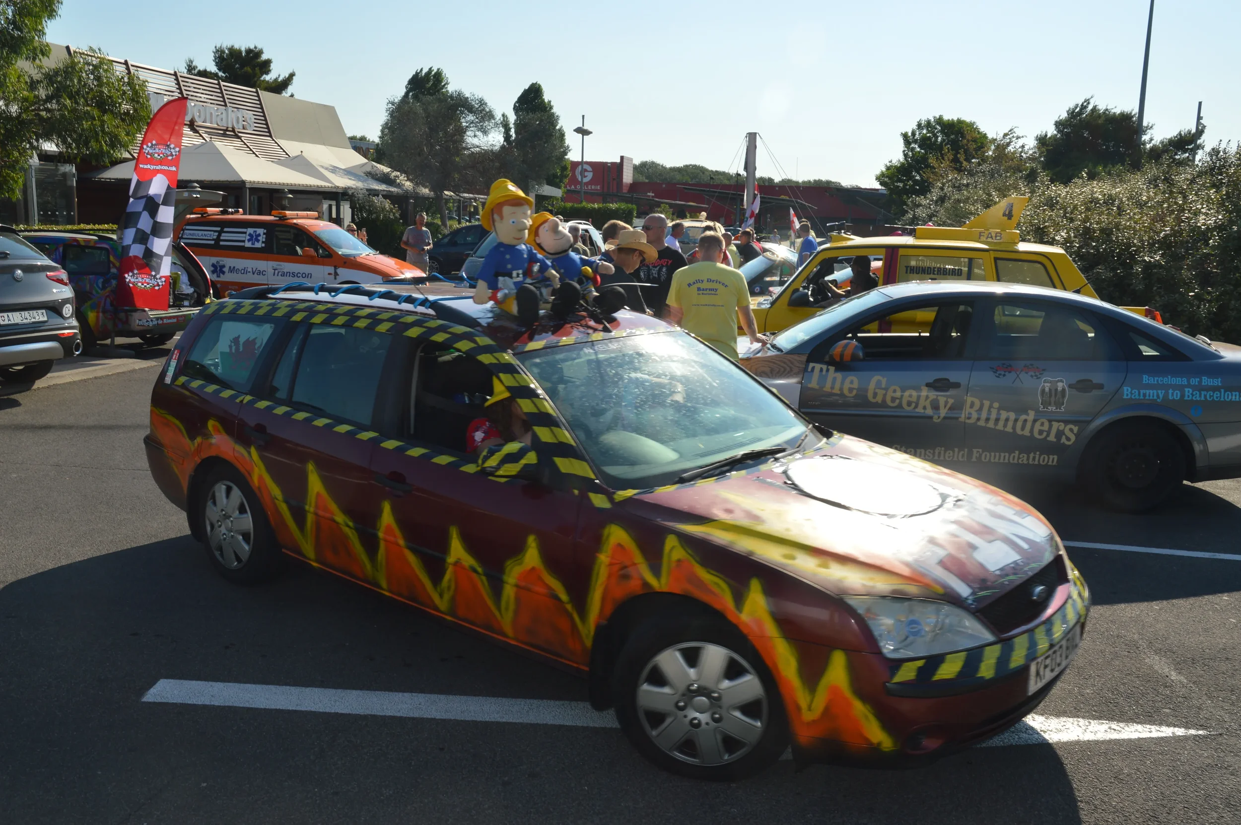 Colorful car decorated with flames and checkered tape, parked among other festival vehicles and emergency vehicles at a gathering or parade.