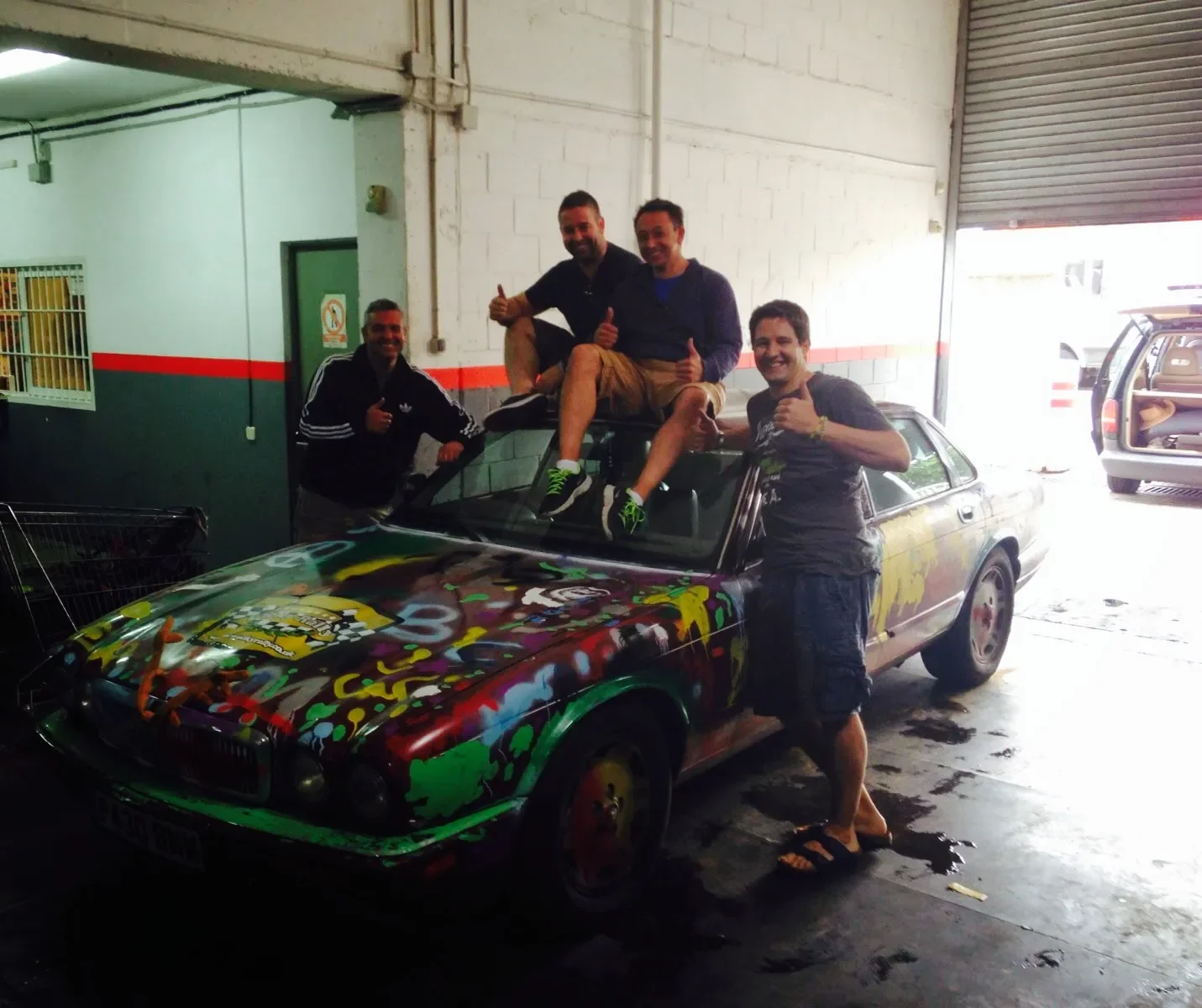 Four men smiling and giving thumbs up pose around and on a colorful, graffiti-painted sports car inside a garage.