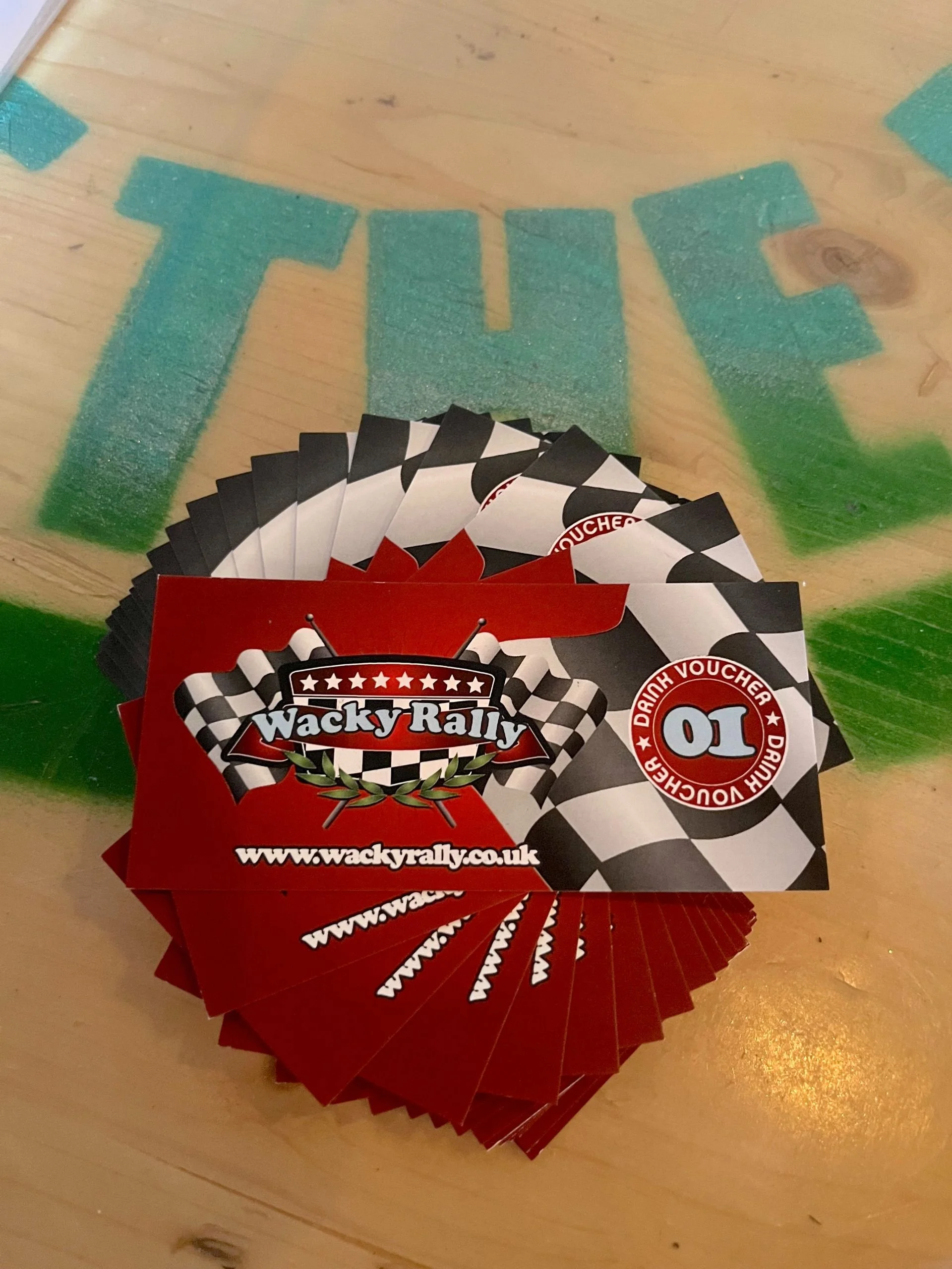 A stack of red and black Wacky Rally gift cards with checkered flags and a red ticket with the number 10 and 'Driver Voucher' text, placed on a table with a green and blue painted word 'THE' in the background.