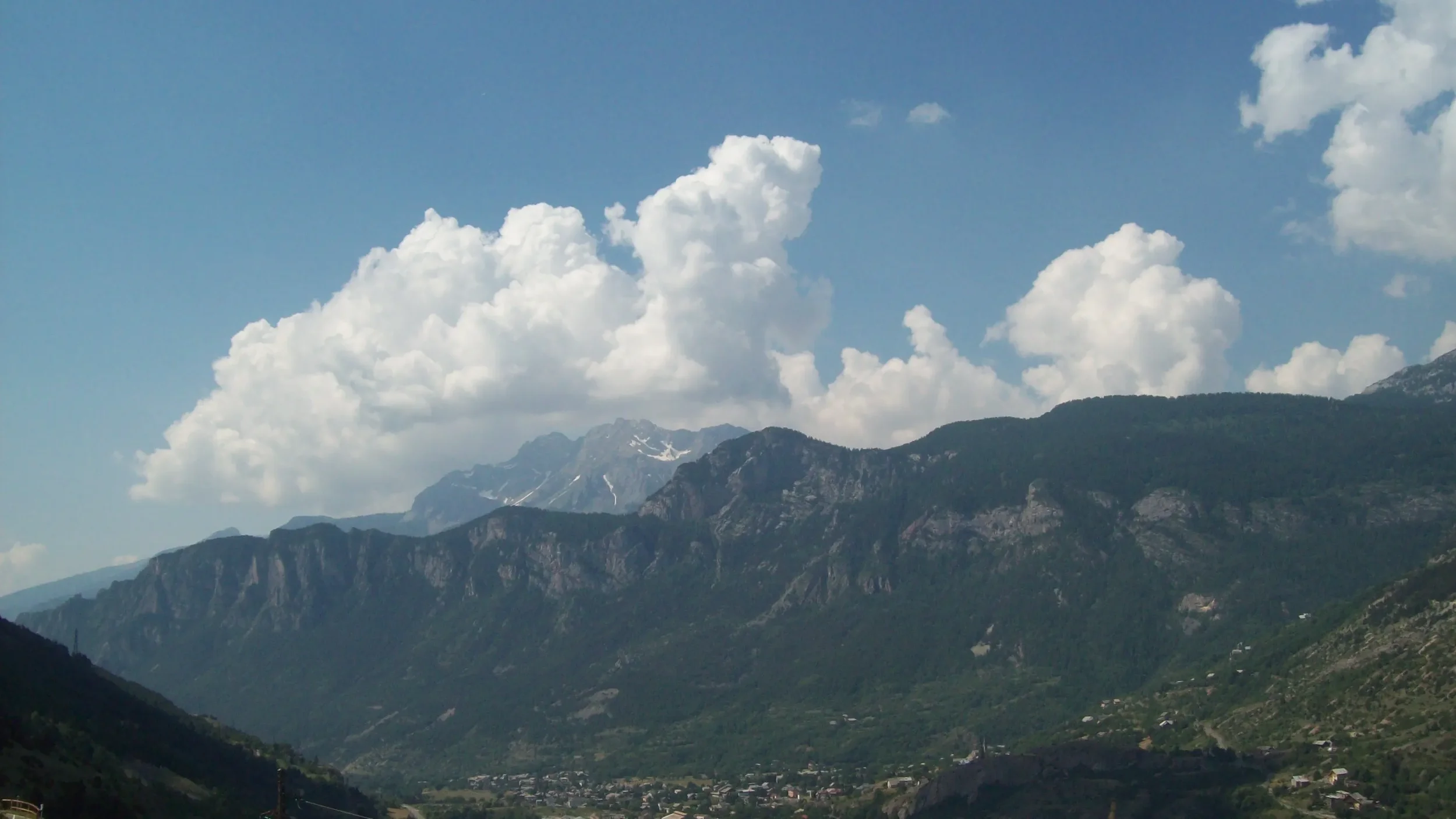 Mountain range with green slopes, cloudy sky with large white clouds, some snow visible on distant mountain peaks.
