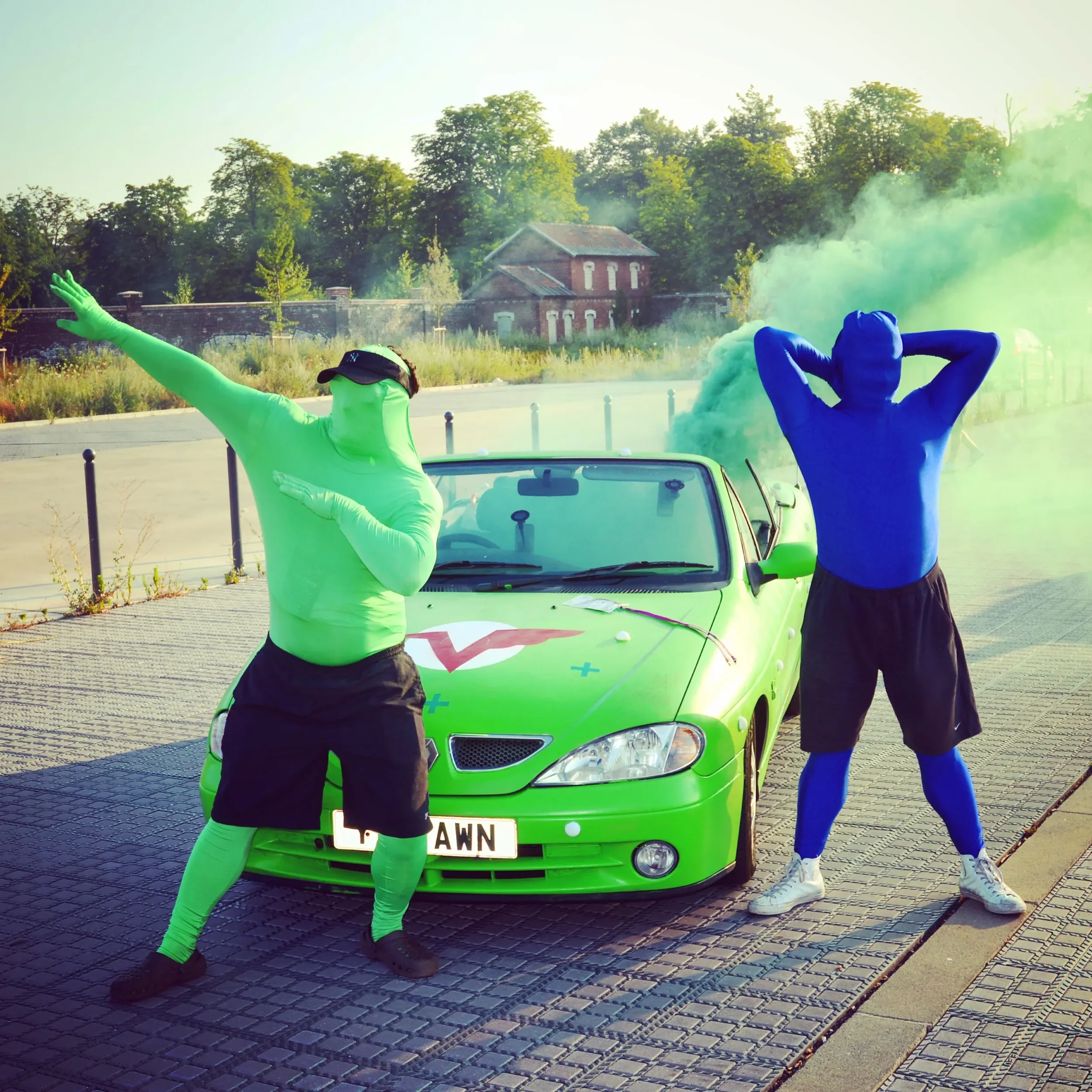 Two people dressed in colorful full-body suits, one green and one blue, posing enthusiastically near a green car with smoke or colored fog in the background, outdoors on a paved area.