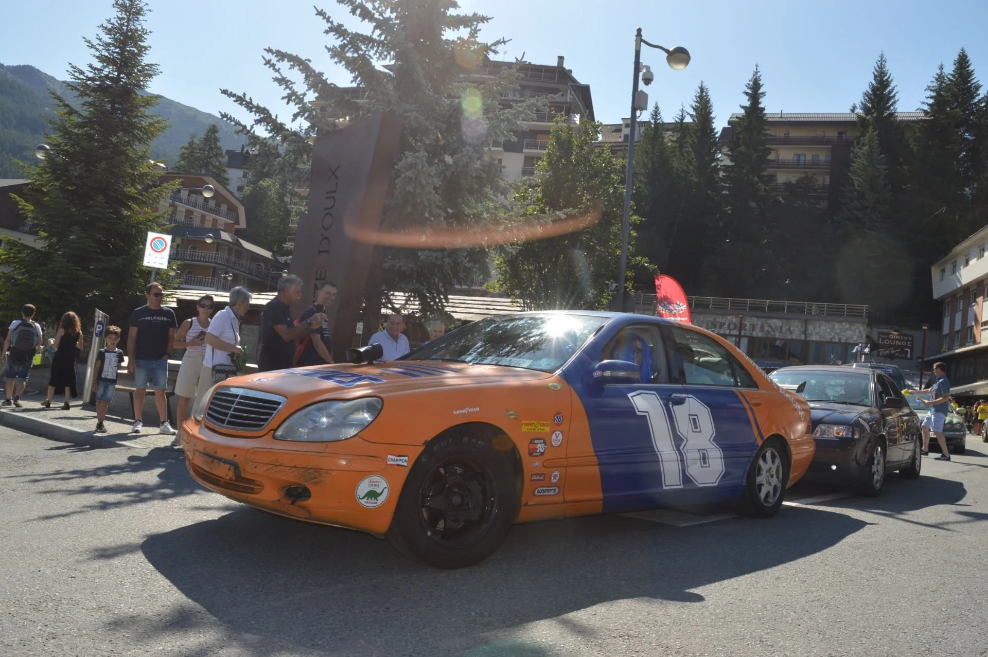 A vintage racing car with the number 18 on its blue and orange body, parked on a street with spectators and other vehicles, in a mountainous area with trees and buildings in the background.