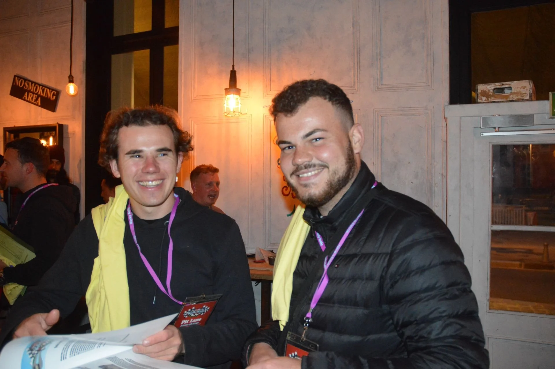 Two smiling young men with lanyards around their necks, standing indoors near a wall with warm lighting. One is holding a paper, and other people are visible in the background.