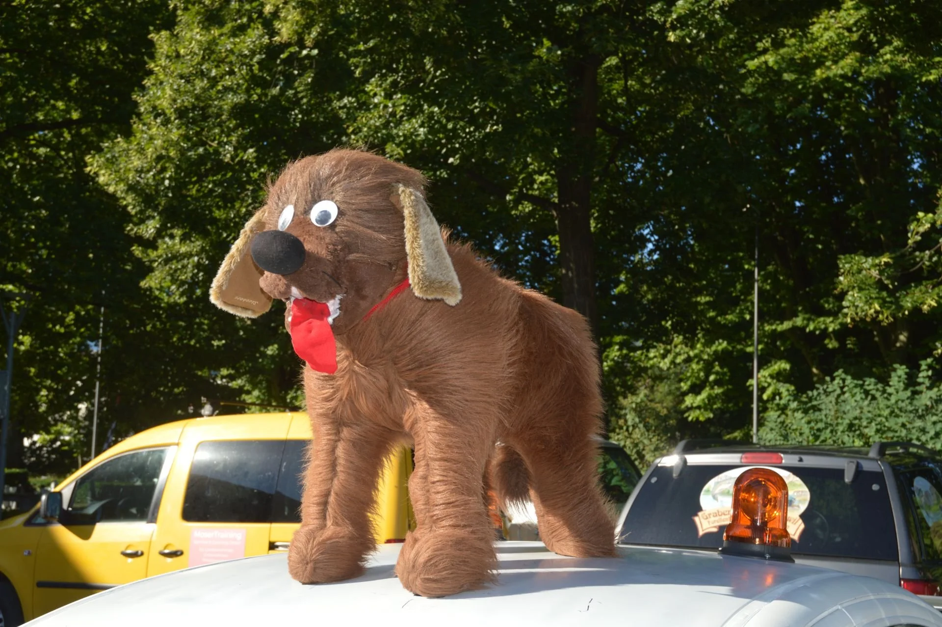 A plush dog costume on top of a police vehicle, with trees and a yellow van in the background.