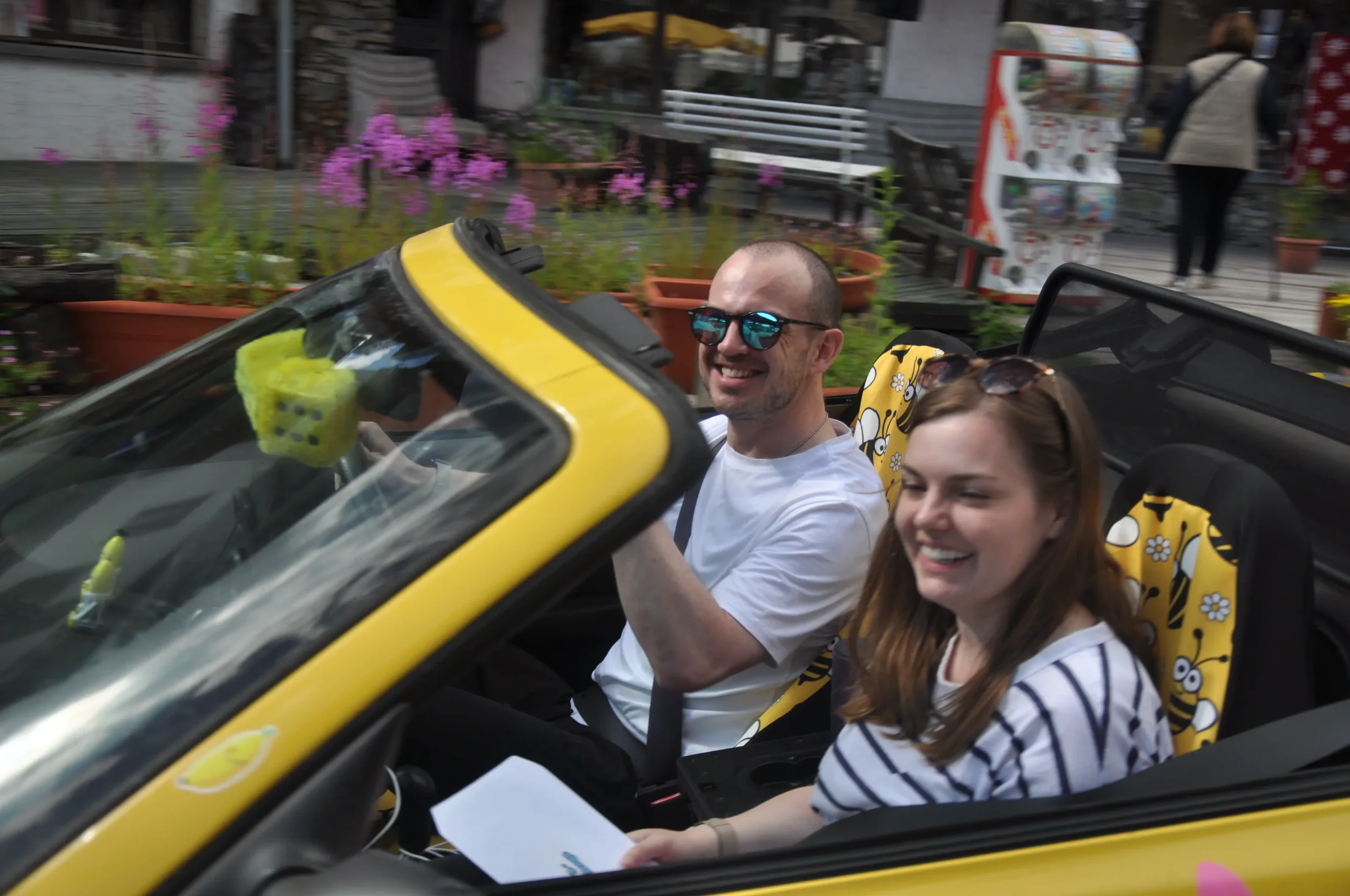 A man and woman sitting in a yellow convertible car, smiling and looking happy. The man is wearing sunglasses and a white t-shirt, and the woman has sunglasses on her head and is wearing a striped shirt. The background shows potted plants, flowers, a