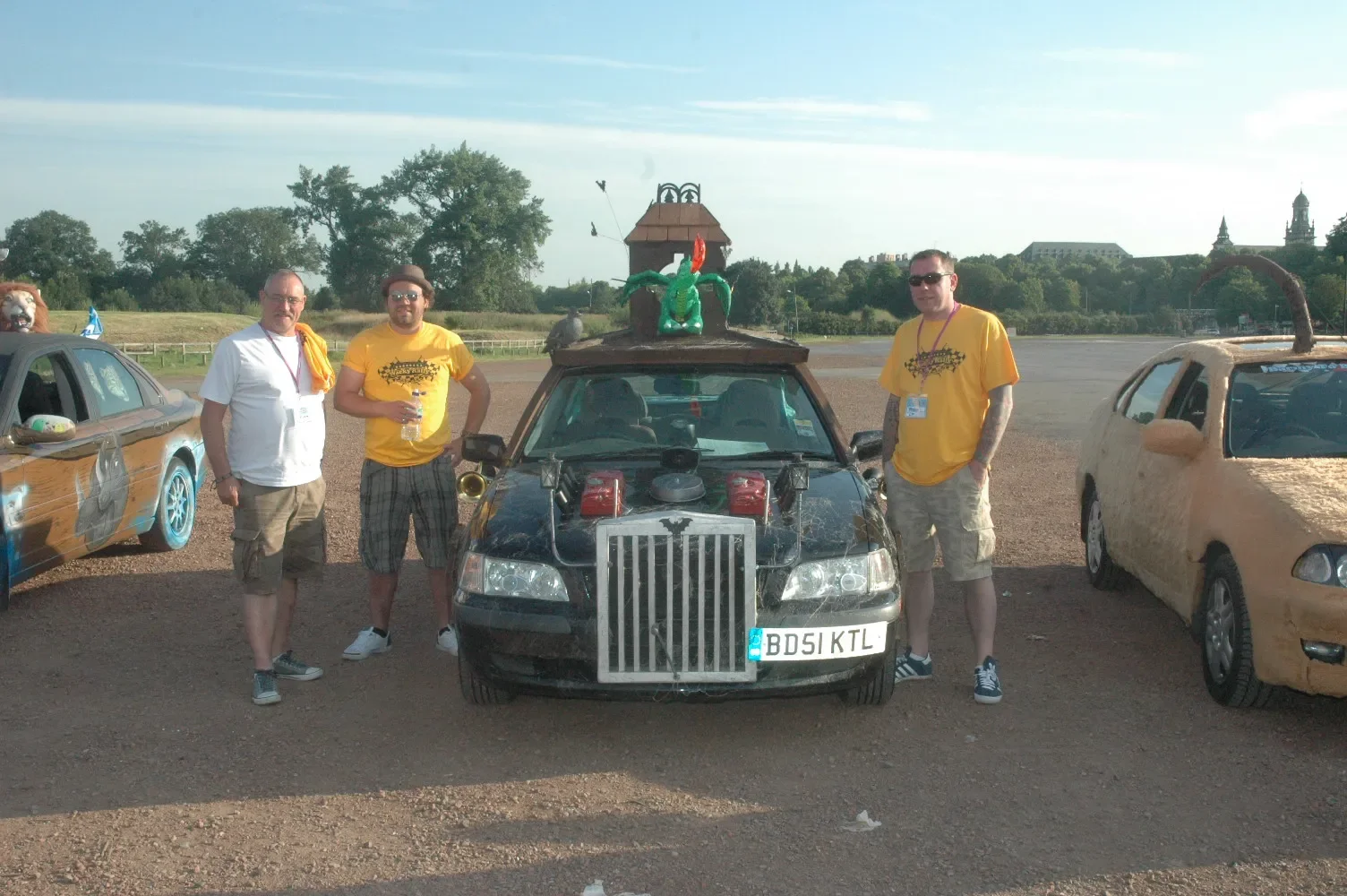 Three men standing in front of a black modified car with a dragon sculpture on top, at a car event with other decorated cars and scenic background.