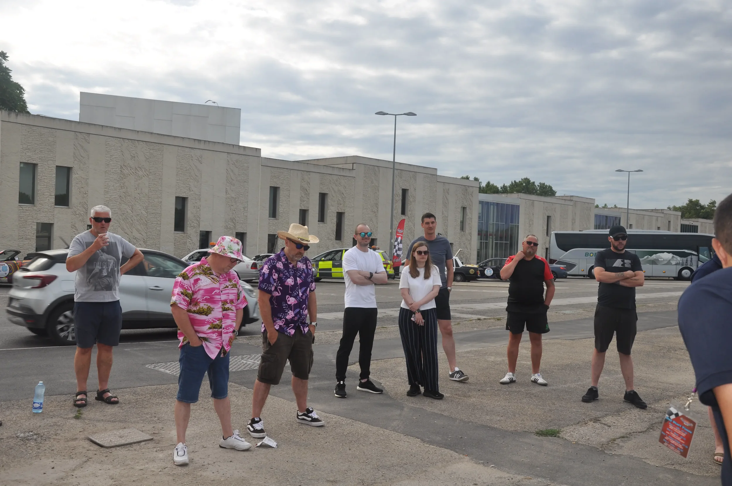 A group of eight people standing outdoors in front of a modern building on a cloudy day, with cars and a bus in the background.