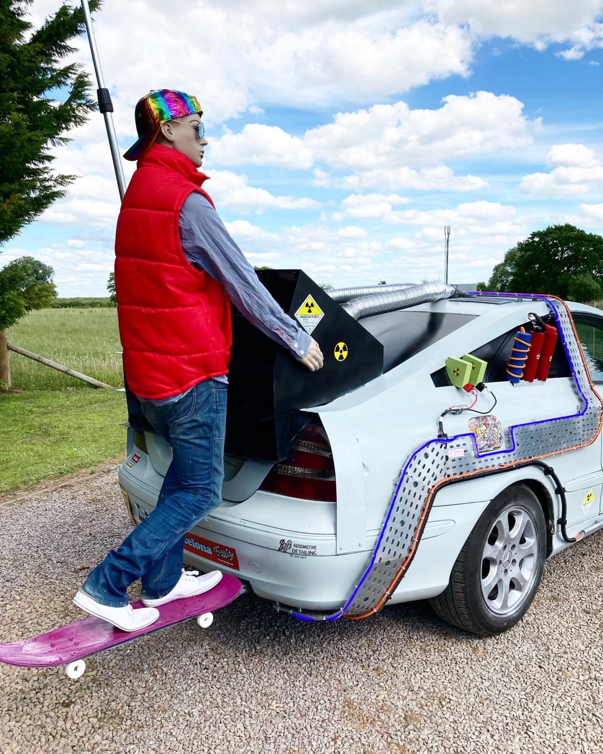A person standing on a pink skateboard behind a modified car that has a large homemade fake nuclear reactor on the trunk, set outdoors on a cloudy day.