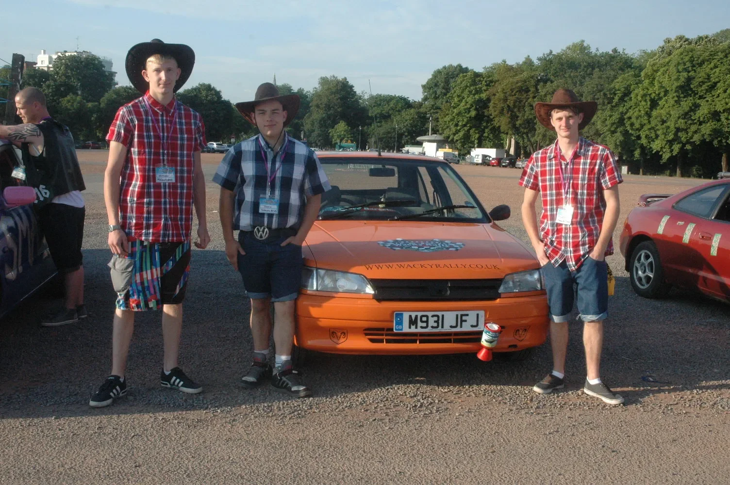 Three young men wearing plaid shirts and cowboy hats standing in front of an orange car at an outdoor event.