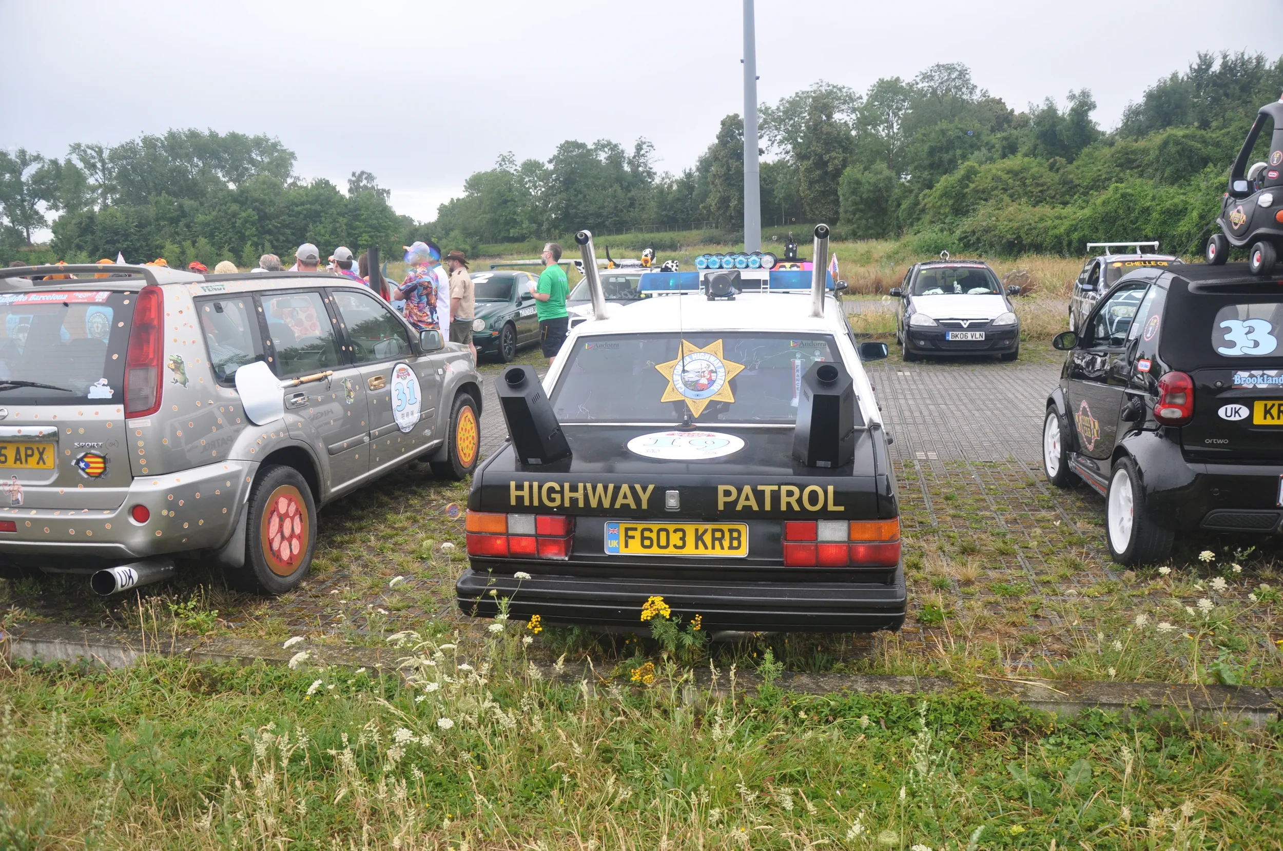 Black highway patrol car with yellow lettering, parked among other vehicles, with a group of people gathered nearby on a cloudy day.