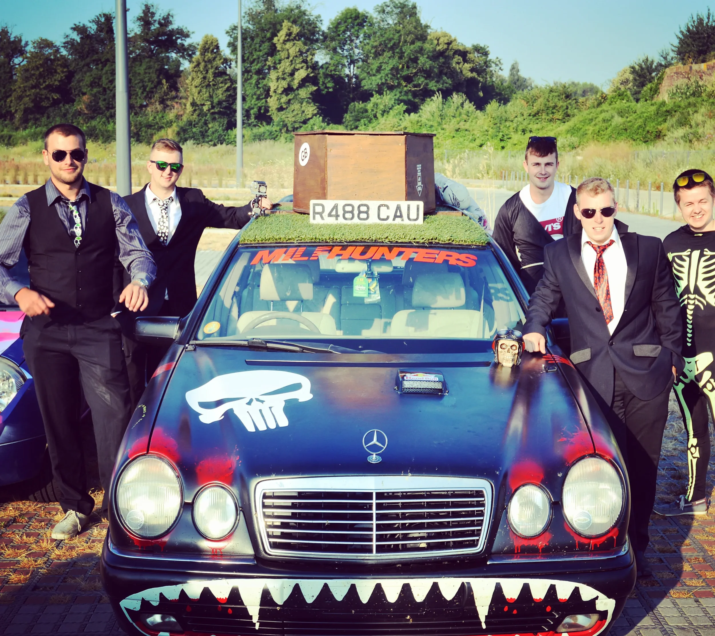 Group of six men in suits and casual clothing standing around a dark-colored Mercedes-Benz with custom paint and decorations, including a skull logo on the hood, a painted mouth with teeth on the front bumper, and a sign on the roof reading "MILF HUN