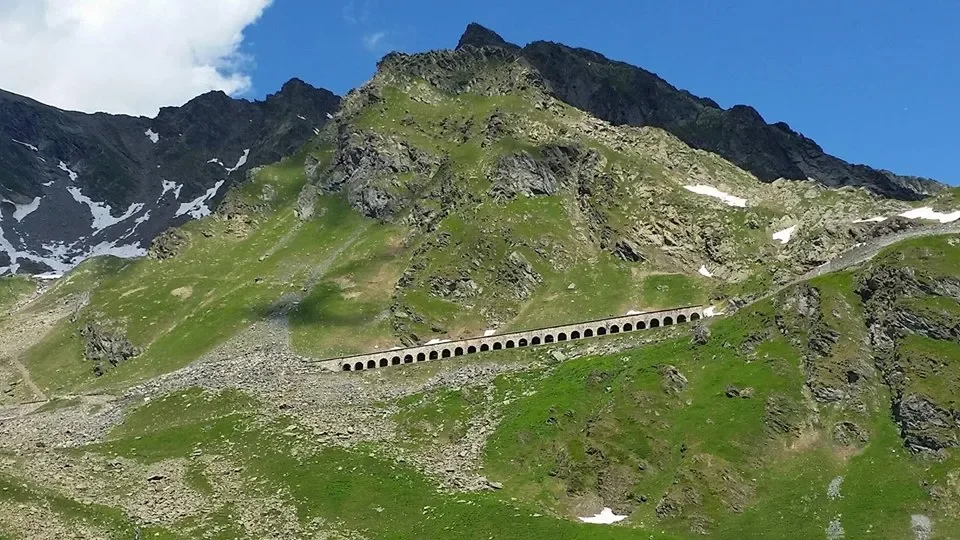 Green mountain landscape with rocky peaks and a tunnel with multiple arches in the hillside, under a blue sky with some clouds.