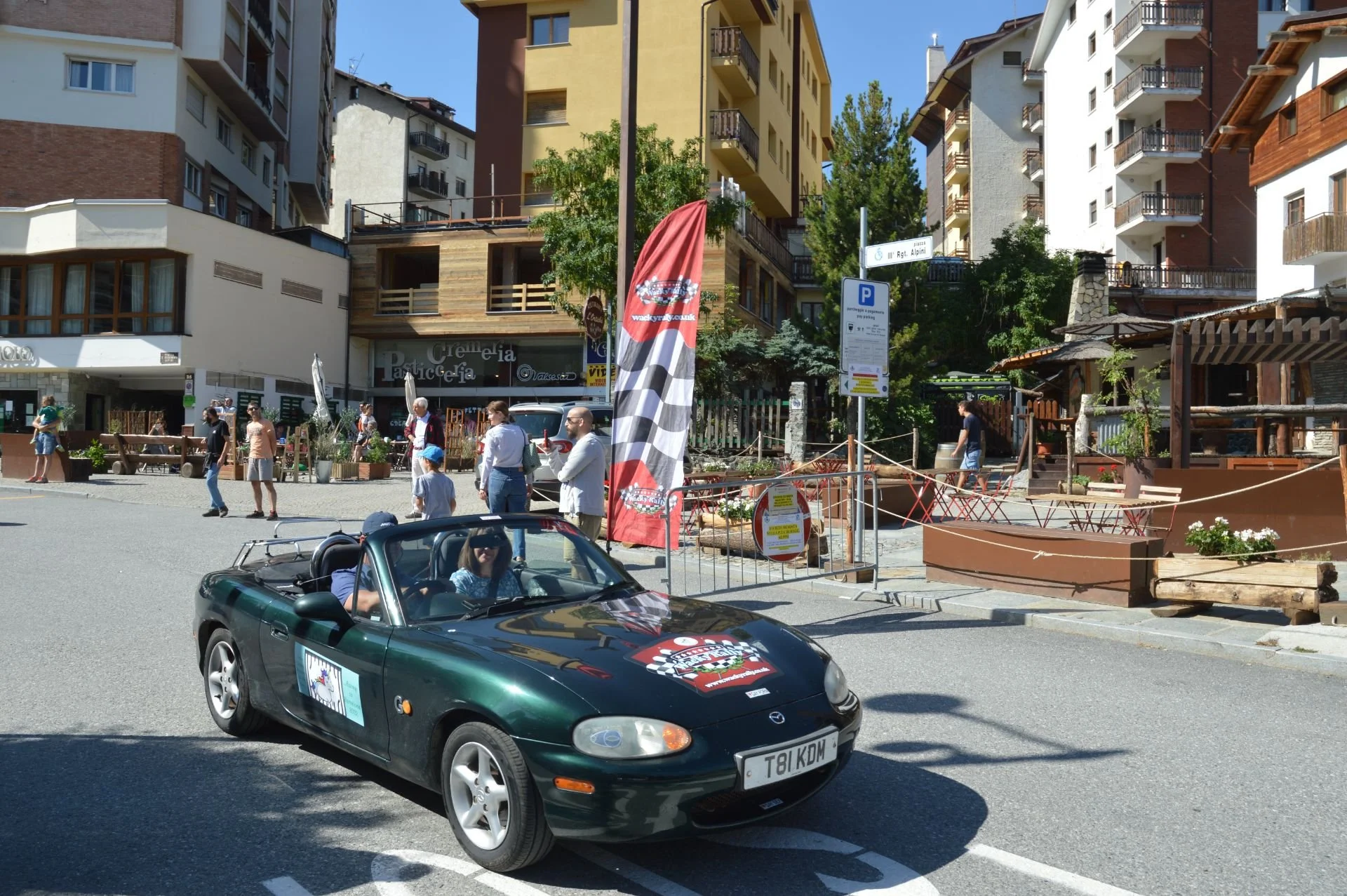 A vintage dark green Mazda MX-5 convertible driving on a city street with several people walking and standing nearby, tall residential buildings, trees, and signage in the background on a sunny day.