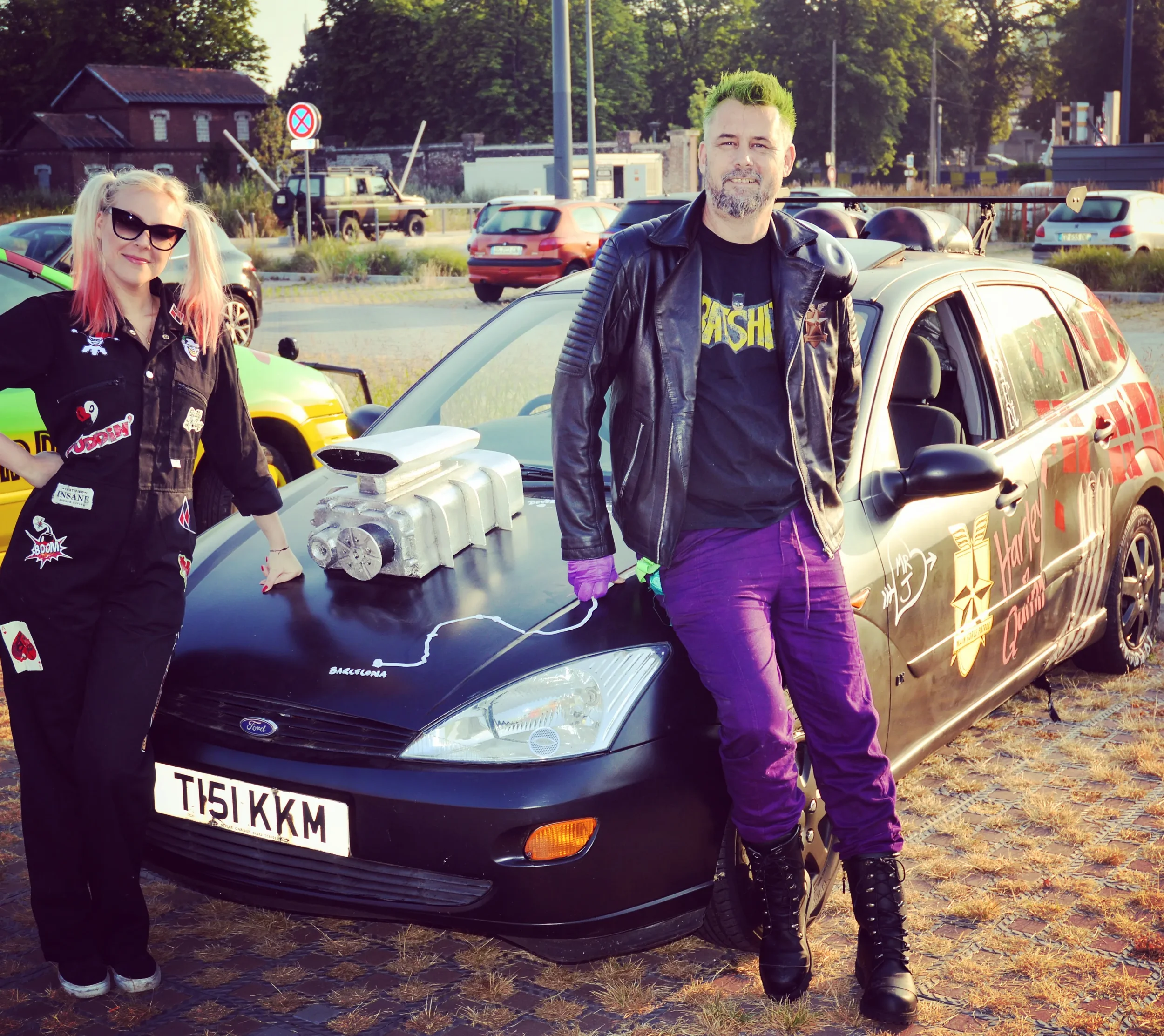 Two people standing next to a black car with a large engine model on hood, in a parking lot. The woman on the left has blonde hair, sunglasses, and is wearing a black jumpsuit adorned with patches. The man on the right has short hair, a beard, and is