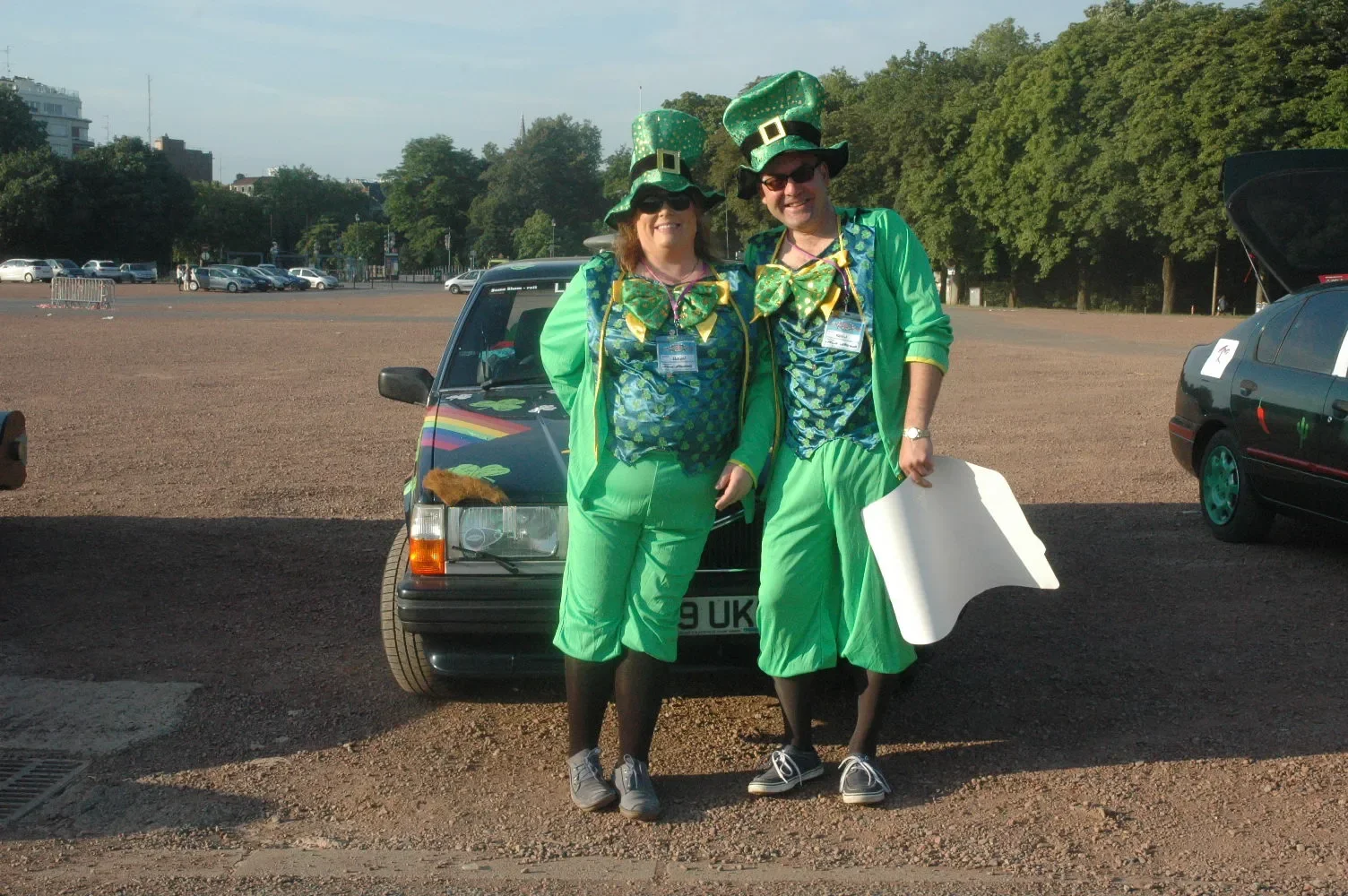 Two people dressed in green leprechaun costumes standing in front of a car, smiling at the camera. They are wearing tall green hats, sunglasses, and have name badges around their necks. One is holding a white poster or board. The scene appears to be 